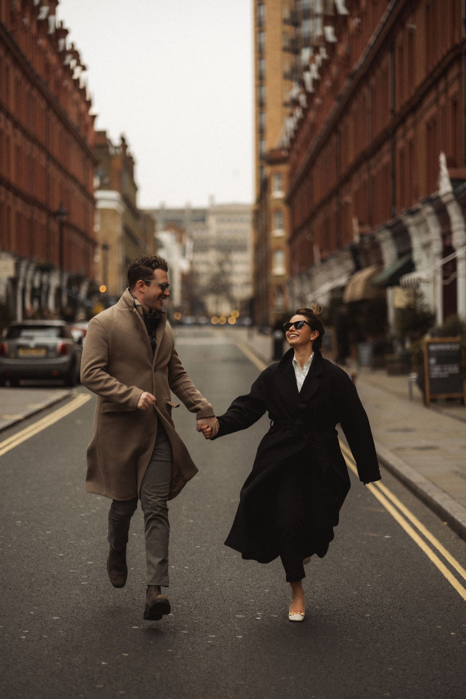 A couple in vintage-style clothing performs playful dance moves on a cobblestone street between brick buildings.