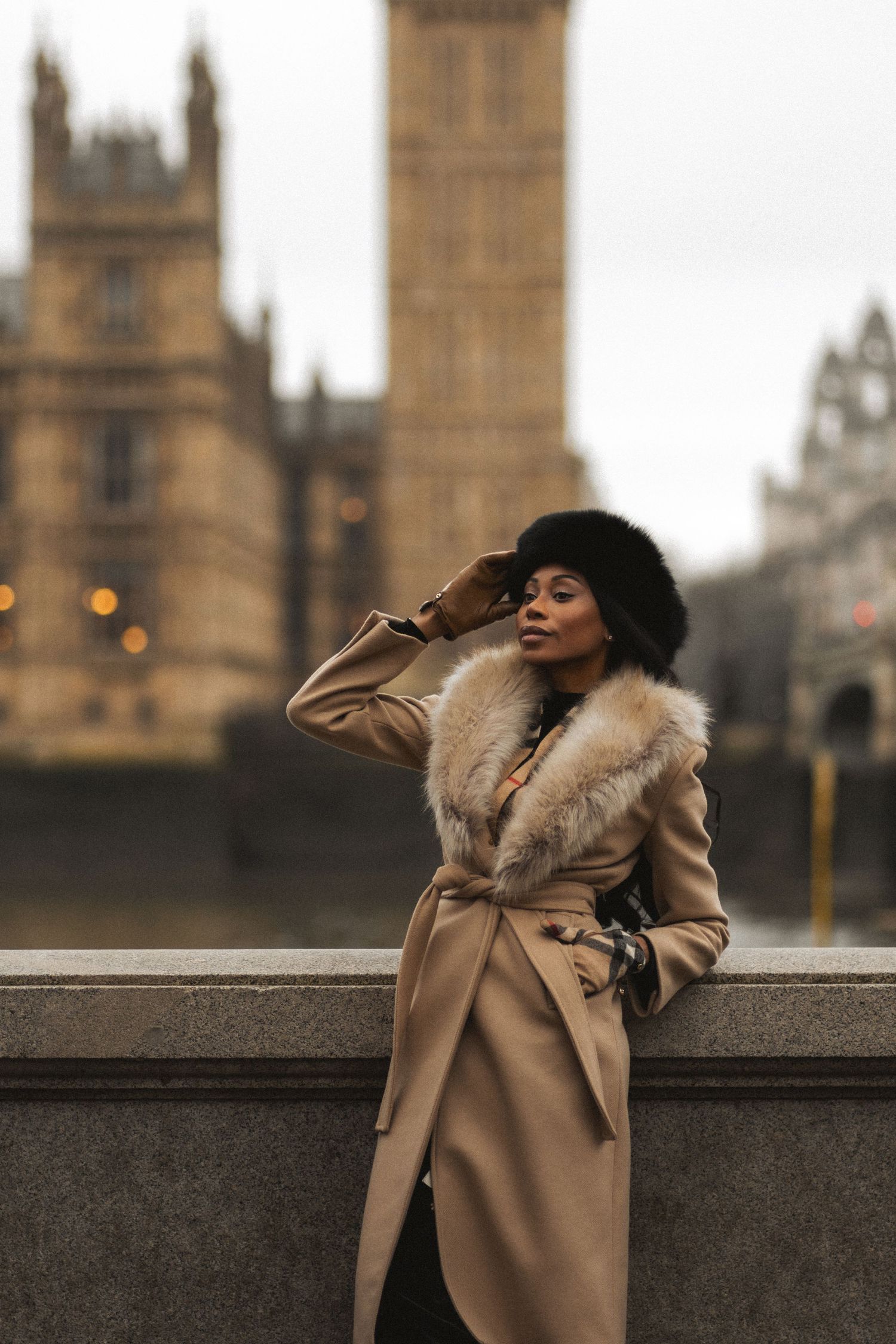 Stylish winter fashion shot featuring a camel coat with fur collar and black hat against London's Houses of Parliament backdrop.
