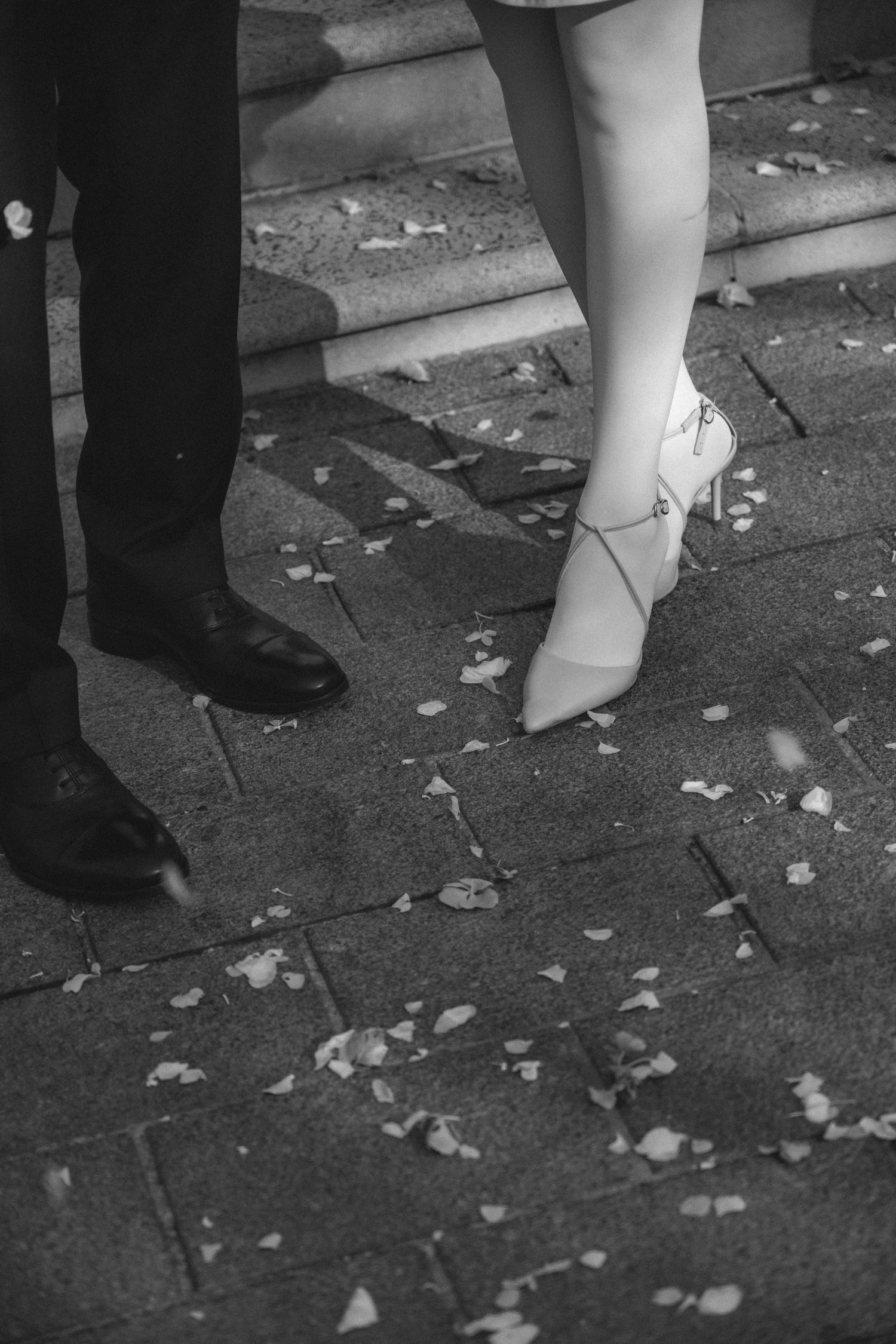 Black and white photograph showing feet of wedding guests standing on ground covered in scattered rice confetti.