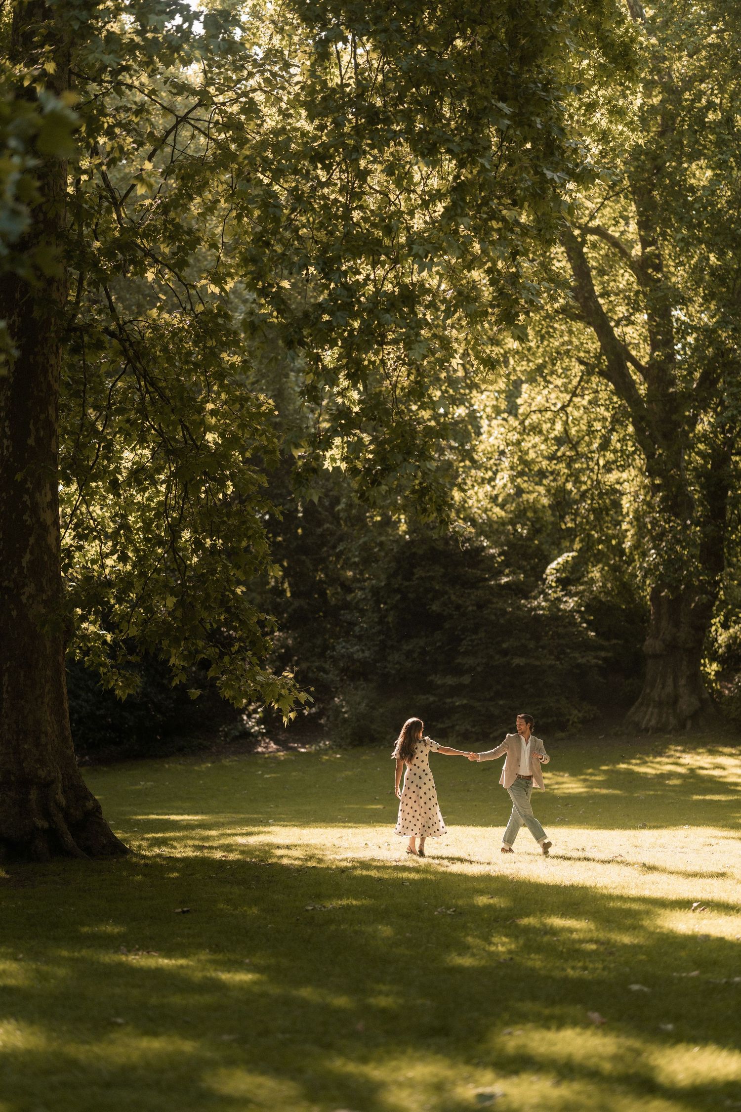Couple dancing hand in hand in a sunlit park surrounded by lush green trees on a warm summer day.
