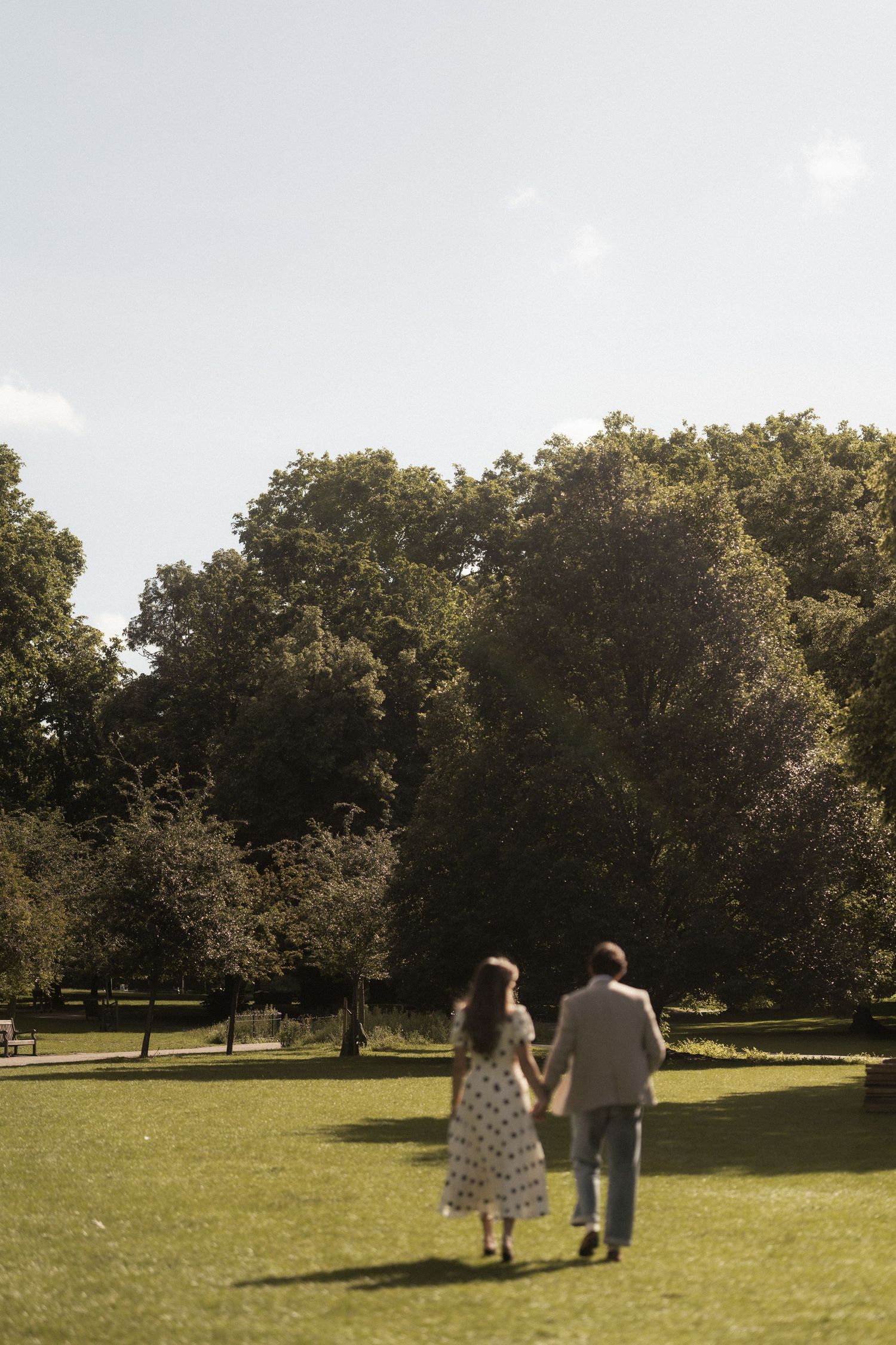 Couple holding hands walking through a lush green park surrounded by tall trees on a sunny day.