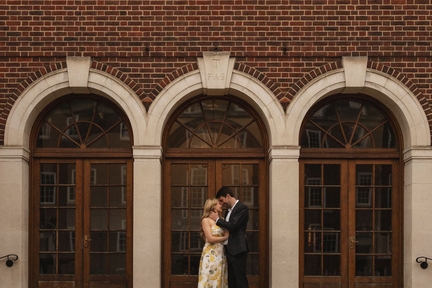 A couple poses in front of an elegant brick building with arched windows and white stone architectural details.