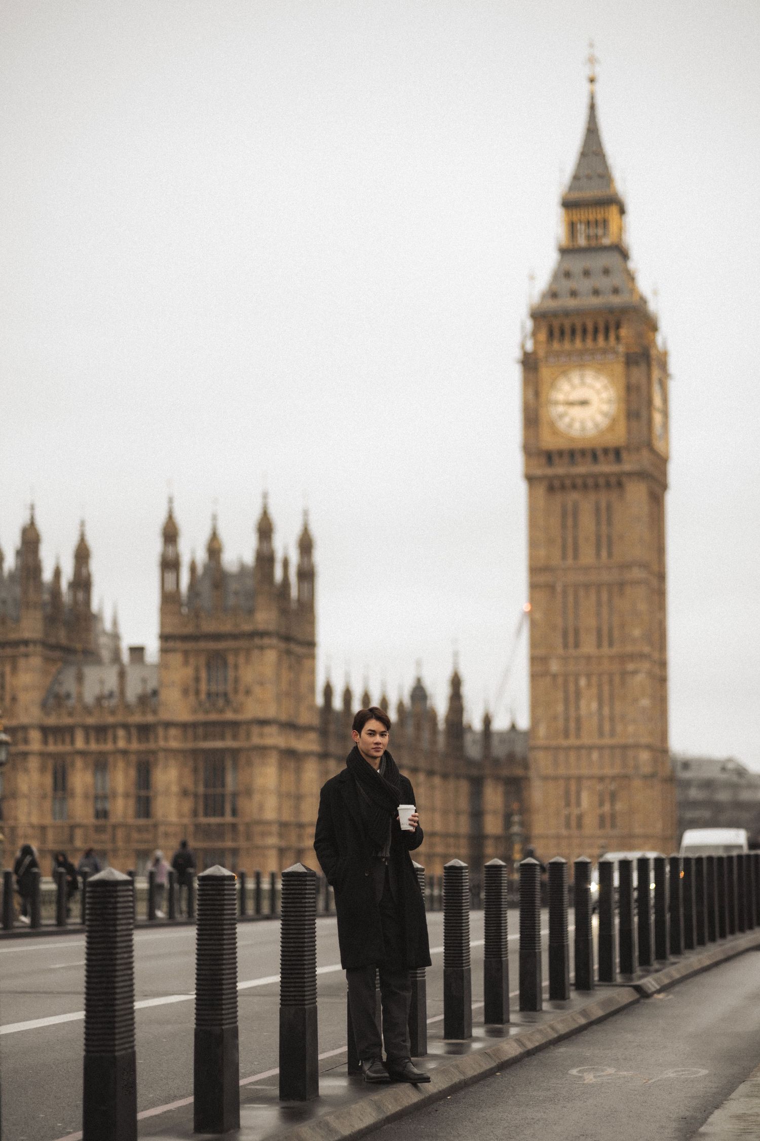 A person in a dark coat stands on Westminster Bridge with Big Ben and Parliament buildings in the background on a cloudy day.