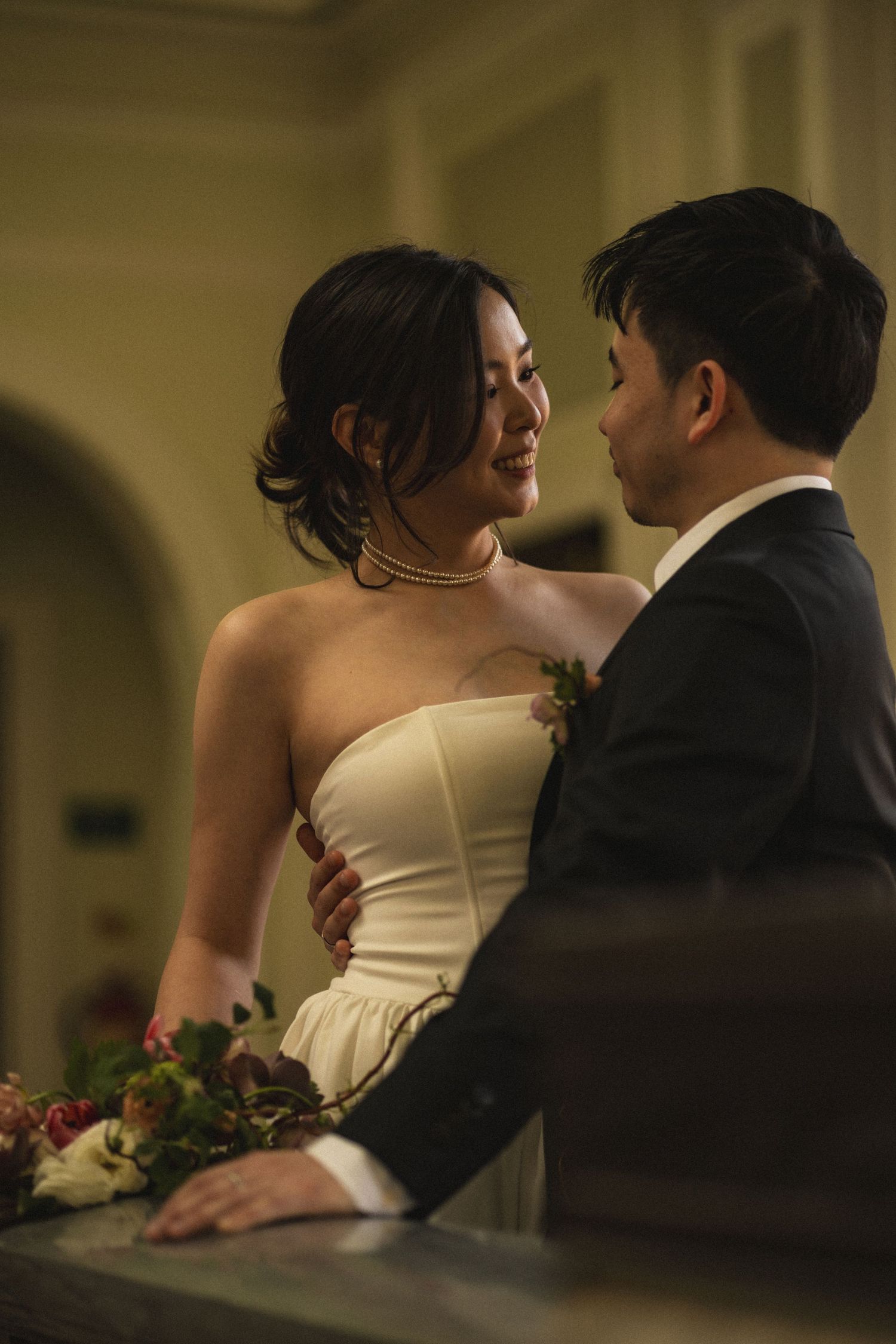 Asian bride and groom share an intimate moment, she in a strapless gown, he in a dark suit, holding a floral bouquet.