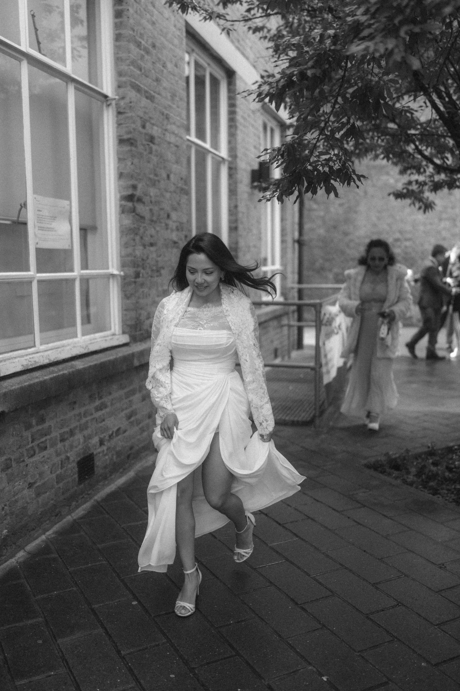 Bride in white dress and lace jacket walks on brick path outside building, black and white wedding photo.