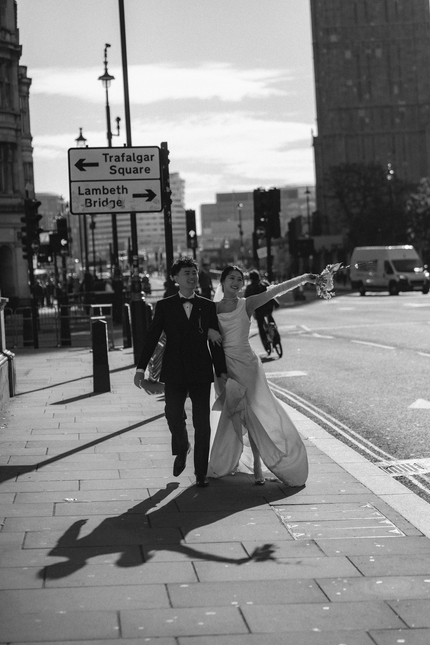 Black and white photo of a bride and groom walking near Trafalgar Square and Lambeth Bridge signs in London.