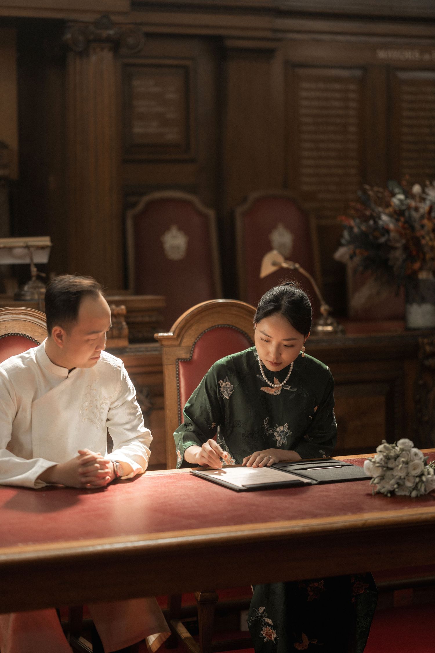Couple in traditional Vietnamese attire signing wedding documents at an ornate wooden table in an elegant venue.