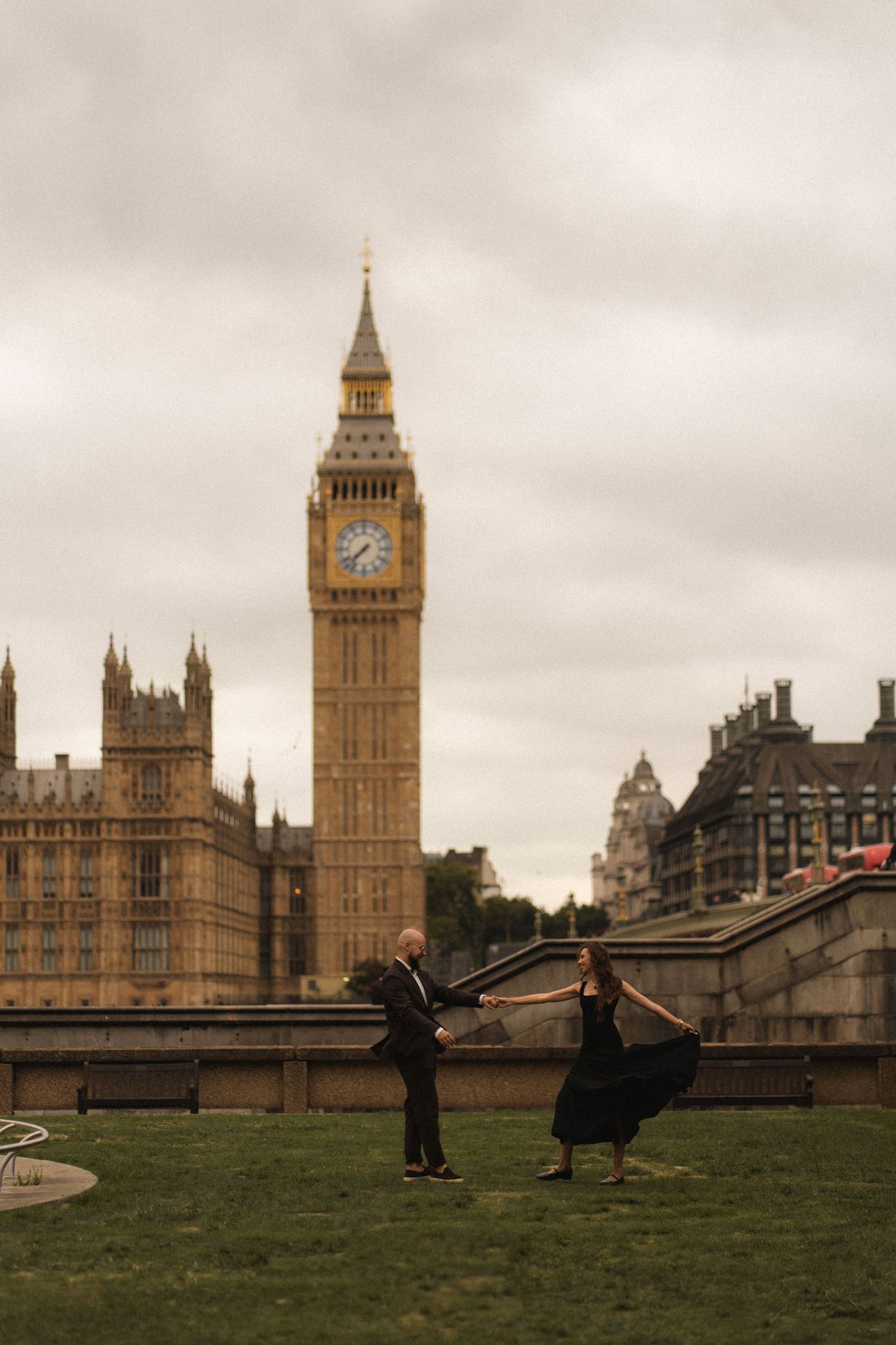 Two people dance playfully on the grass with London's iconic Big Ben and Houses of Parliament as a dramatic backdrop.
