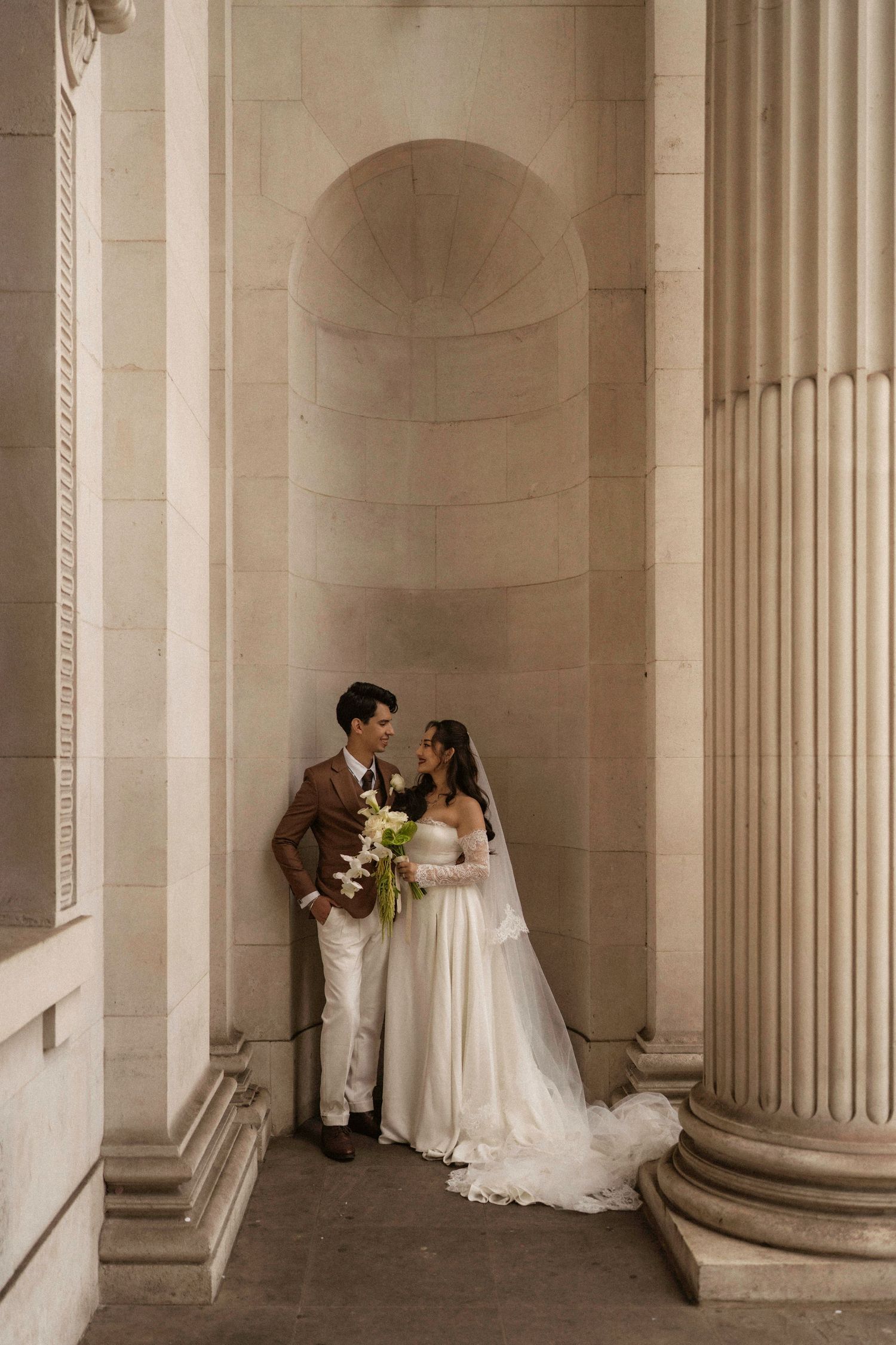 Bride and groom share a romantic moment between elegant stone columns in a classic architectural setting.