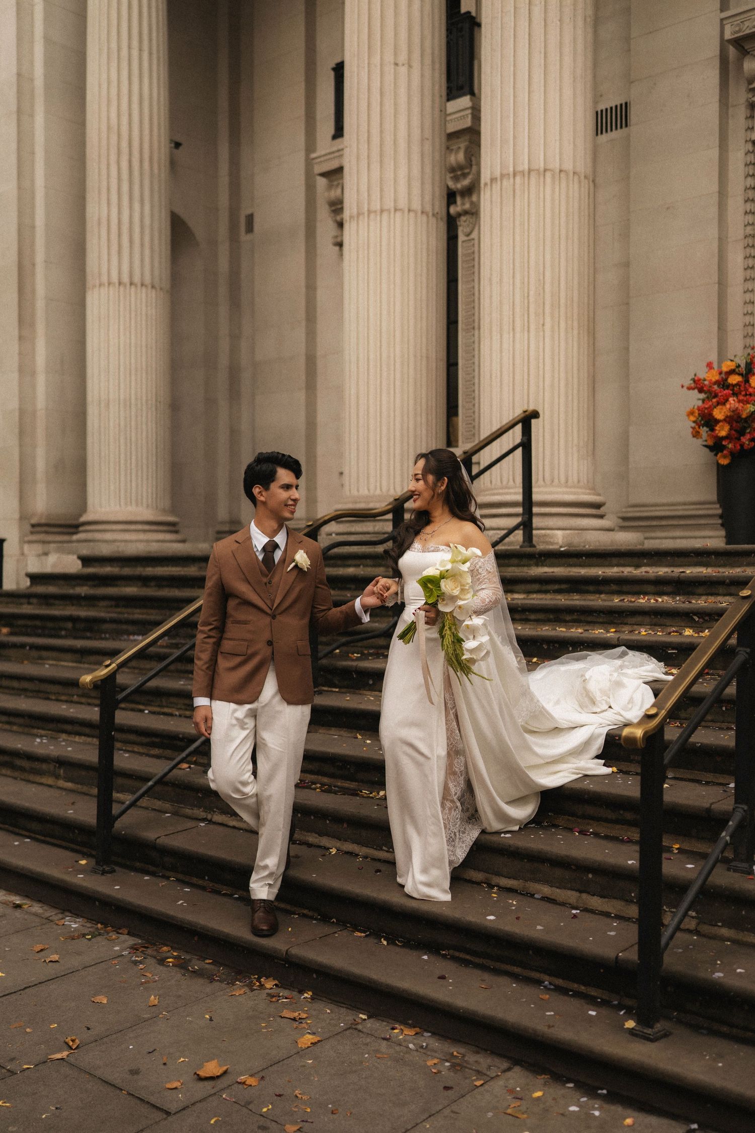 Bride and groom hold hands on grand building steps, she in white gown with bouquet, he in brown suit.