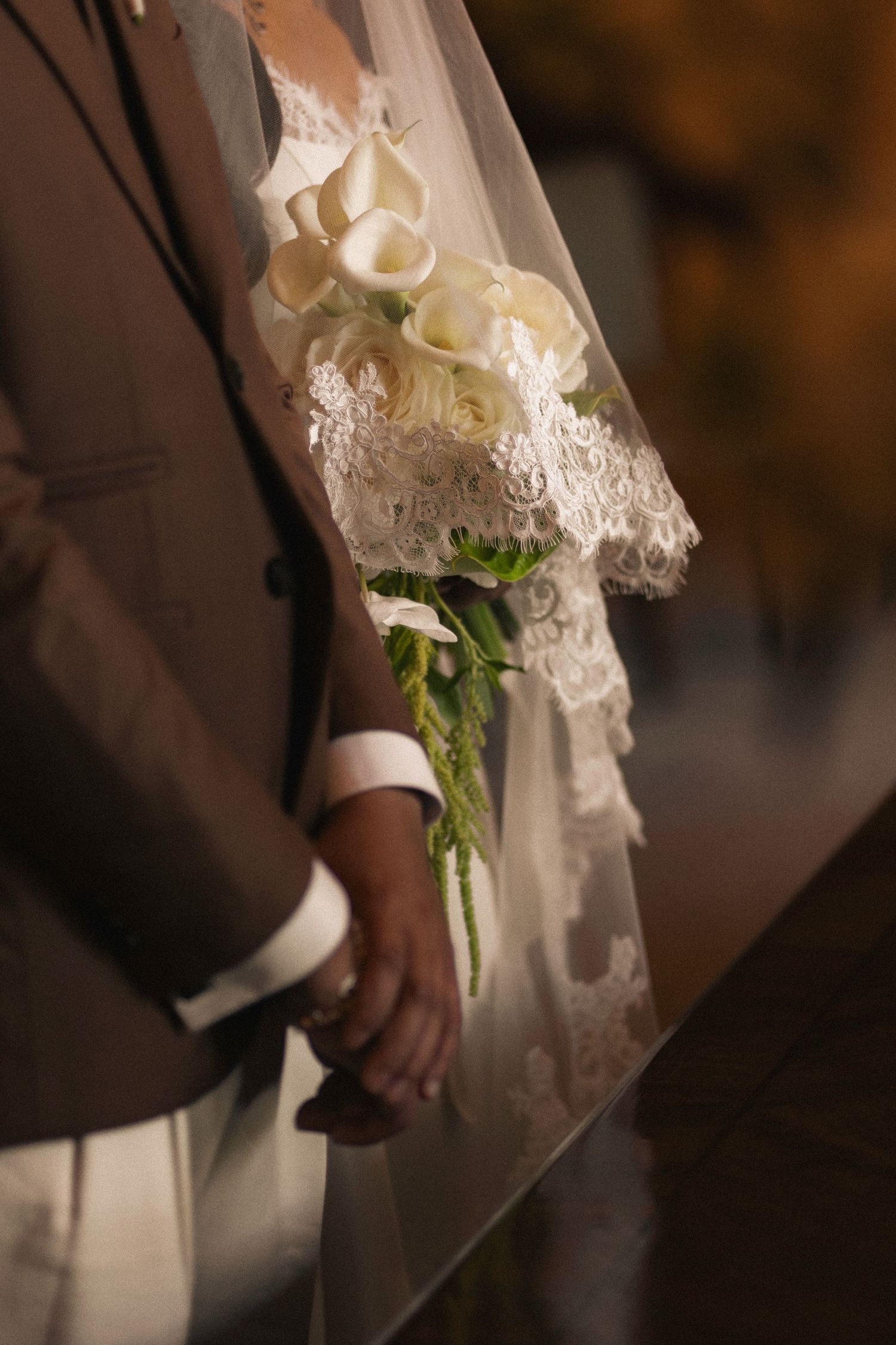 Bride and groom holding white calla lily bouquet, bride in lace wedding dress with veil, groom in brown suit.