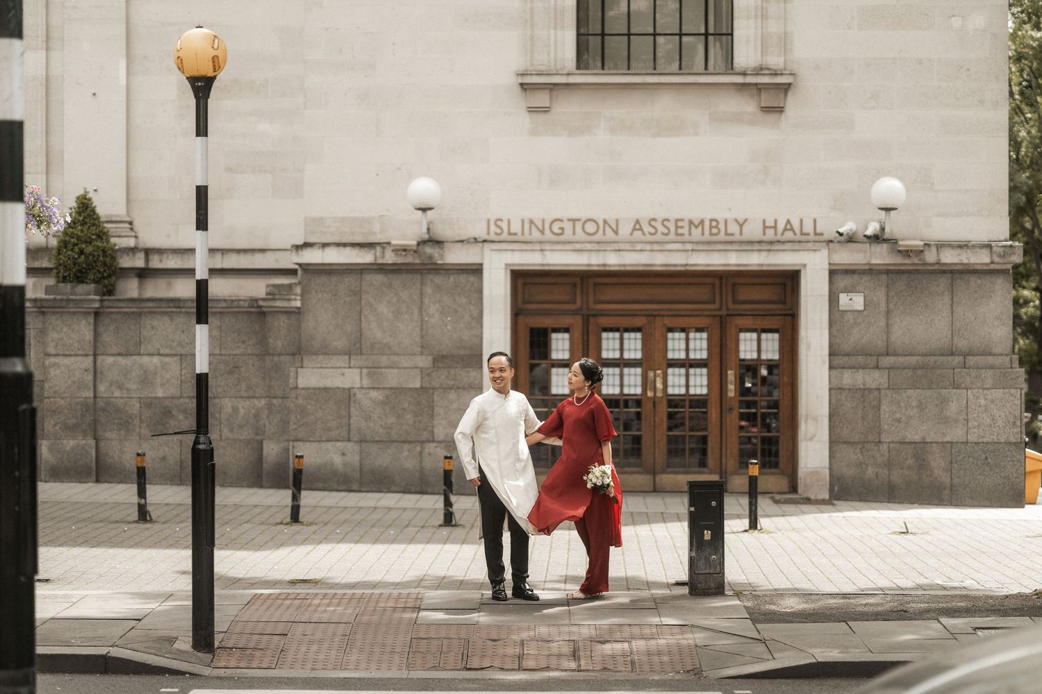 Couple in traditional attire posing outside Islington Assembly Hall on a London street.