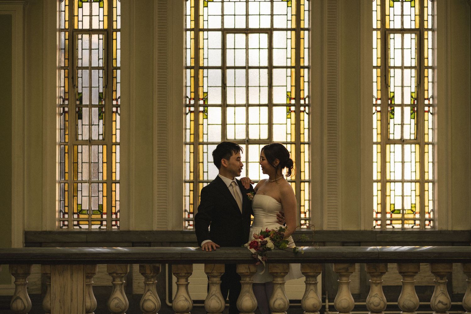 A couple embraces in a grand historic hallway with ornate stained glass windows and stone architecture.