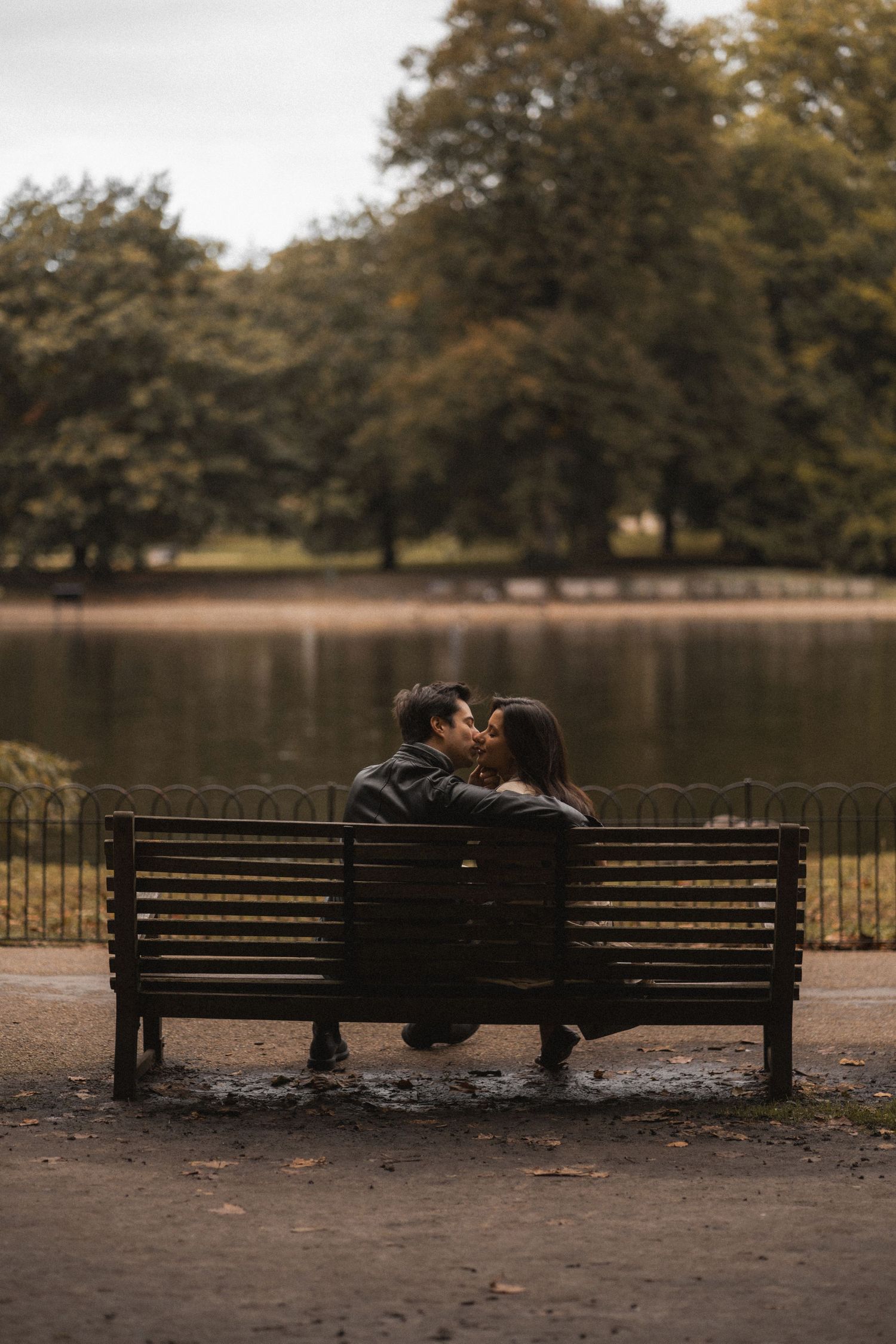 A couple shares an intimate moment on a park bench by a serene lake surrounded by autumn trees.
