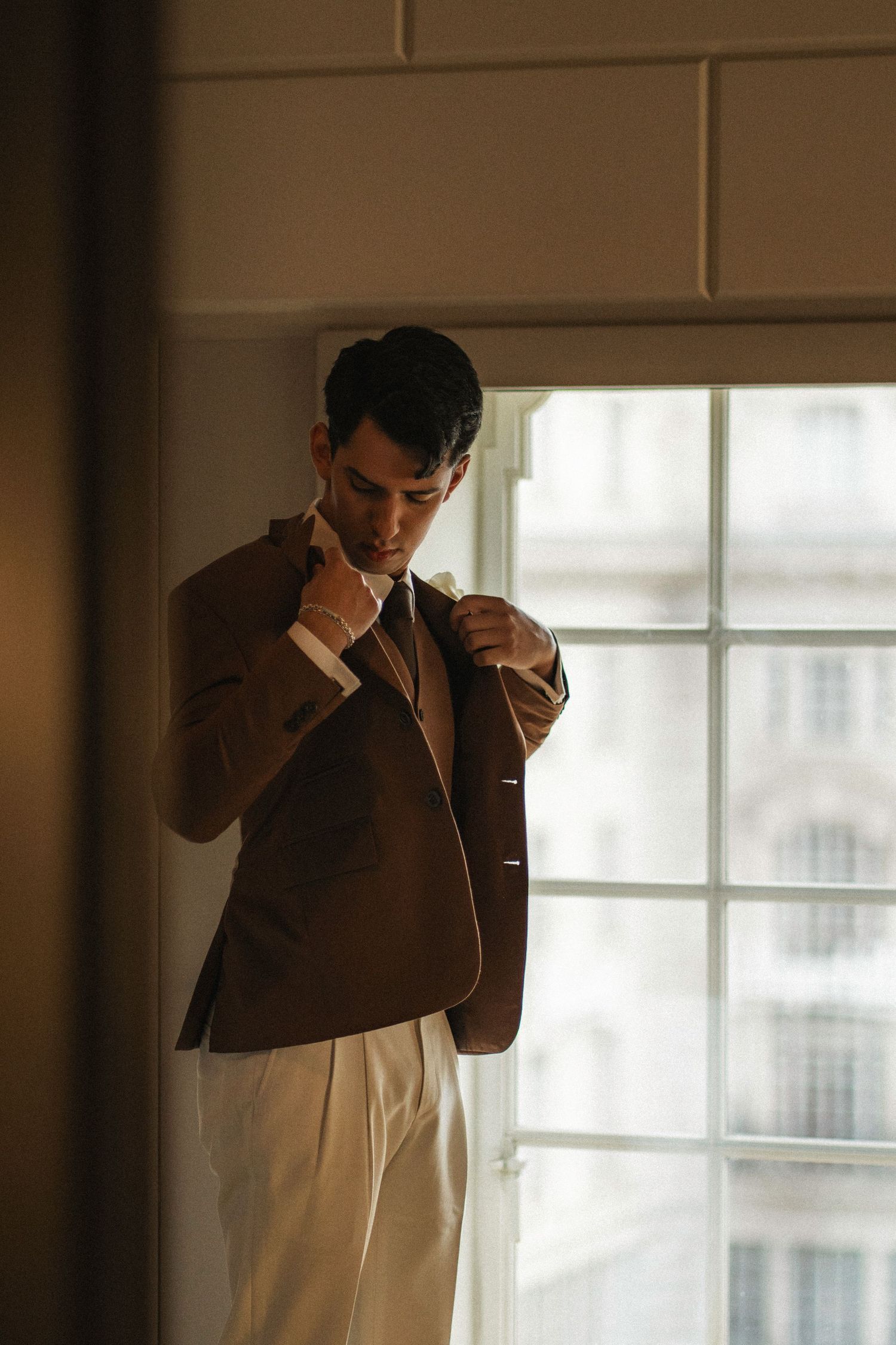 A person in a brown blazer and cream pants stands pensively by a bright window in moody lighting.