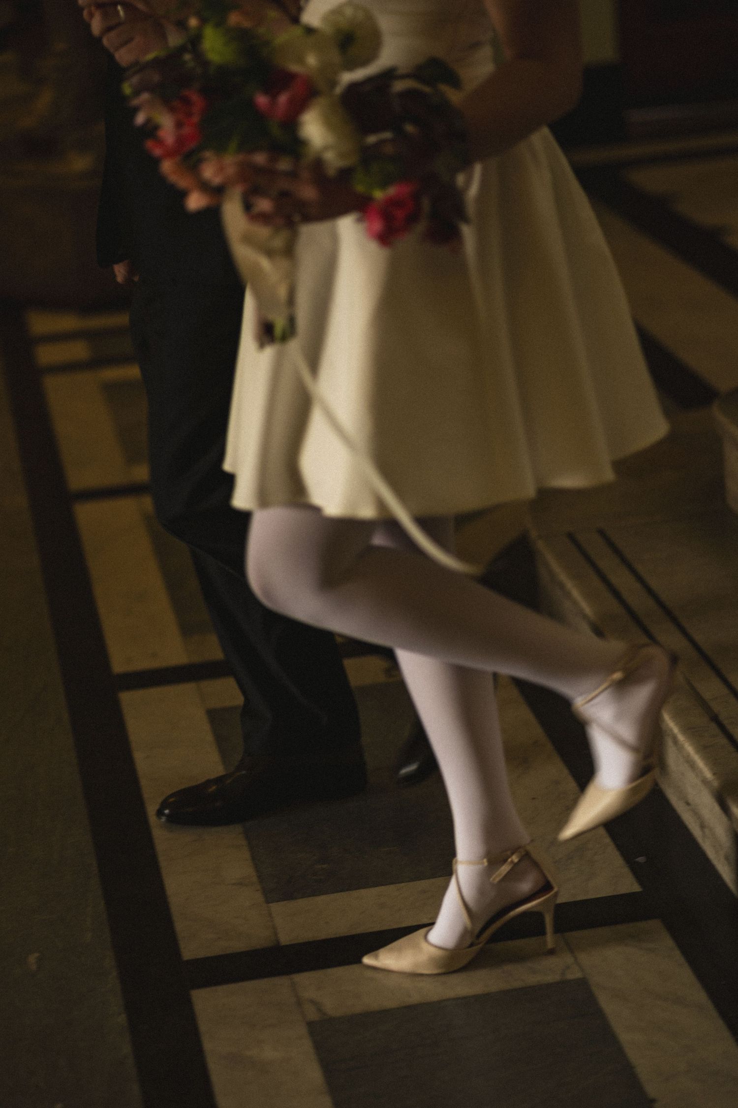 Close-up view of white heels and stockings with a flowing dress hem and floral bouquet visible in dim lighting.