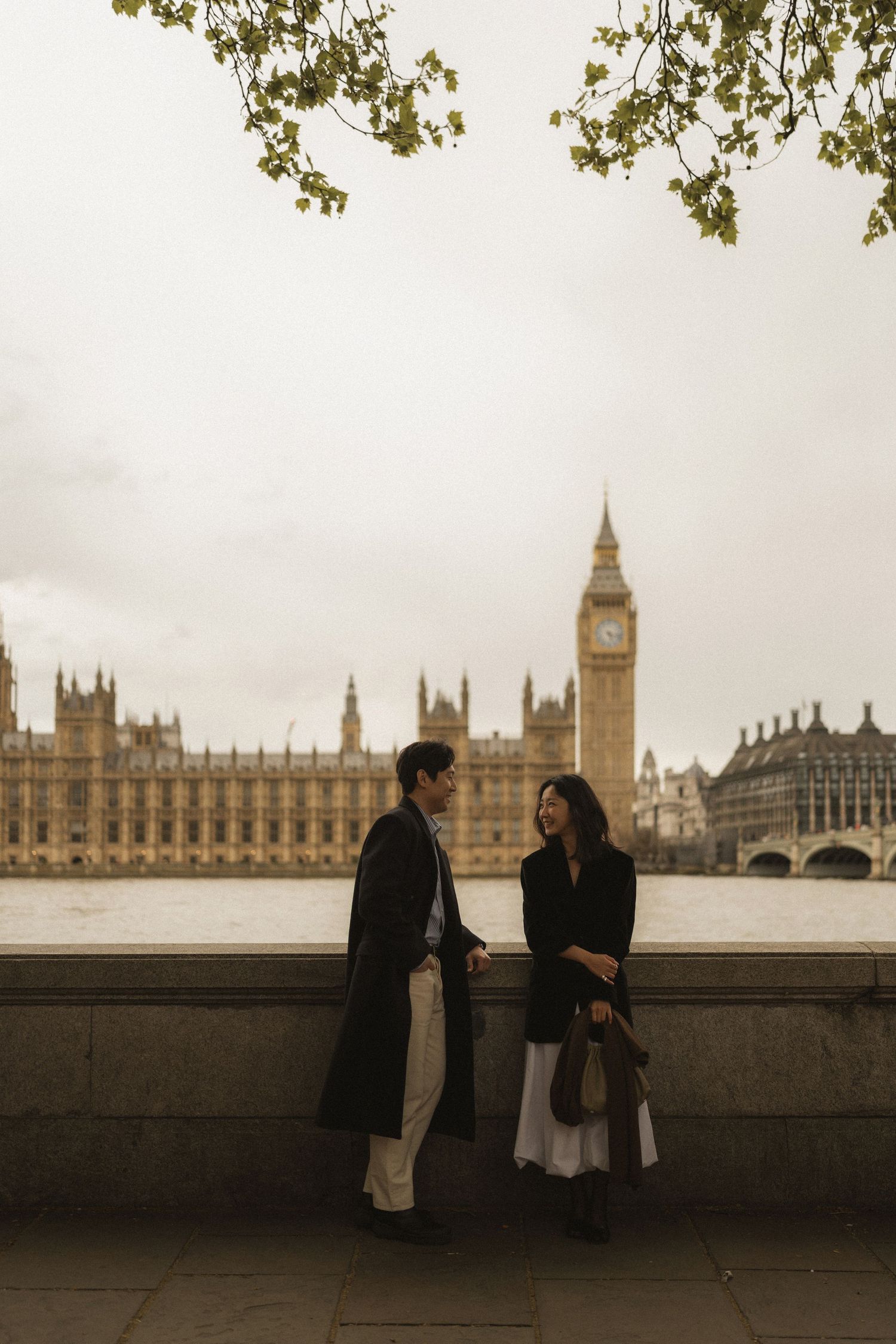 Two people in winter coats stand along Thames River embankment with iconic Big Ben and Parliament buildings in London.