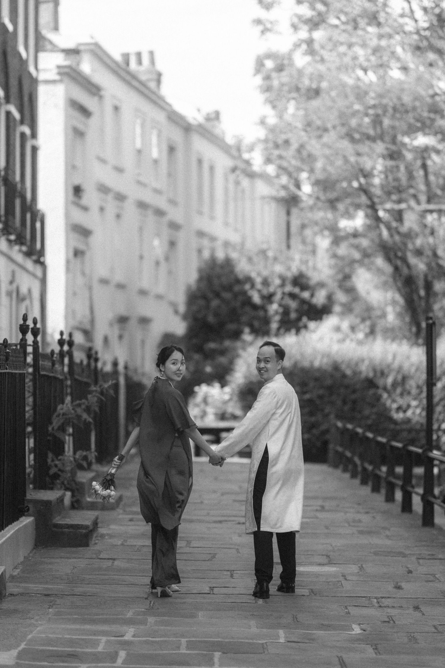 Black and white photo of a couple holding hands on a charming London street, turning to smile at the camera.