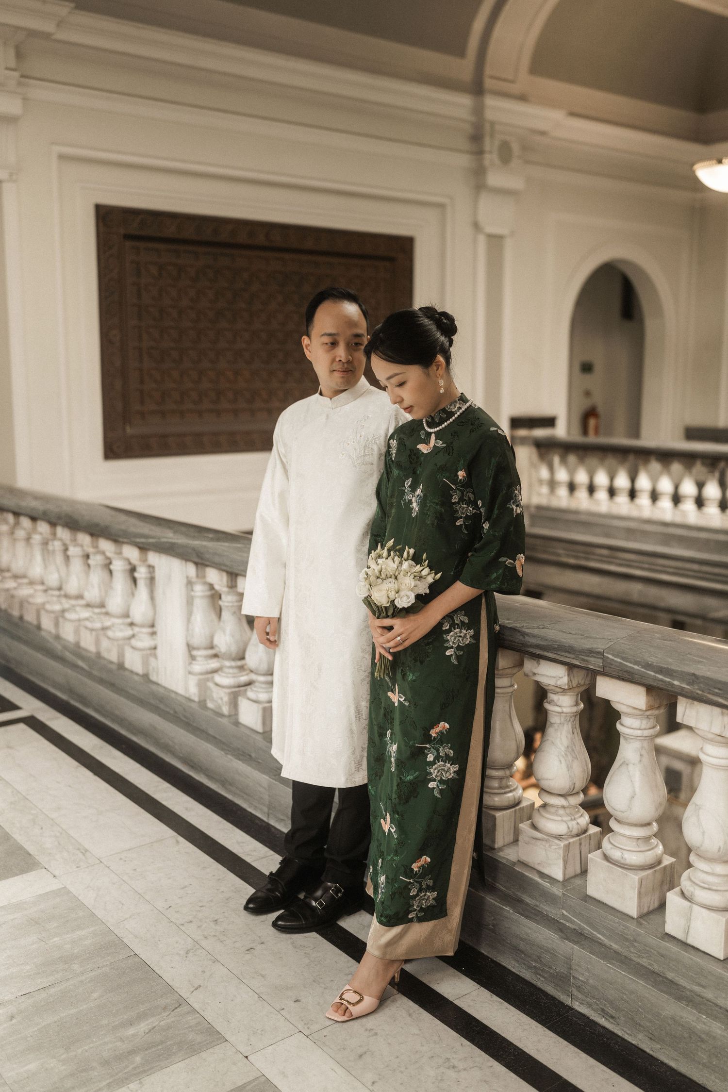 Vietnamese couple in traditional ao dai attire pose elegantly on marble balcony, bride in green floral dress holding white bouquet.