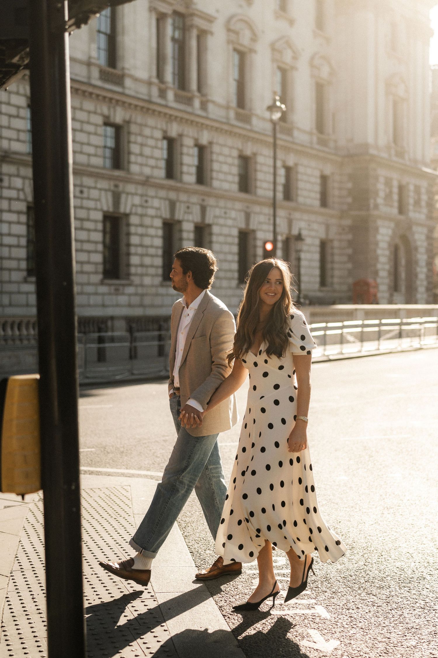 Couple walking on sunny city street, woman in polka dot midi dress and heels, man in beige blazer and jeans.