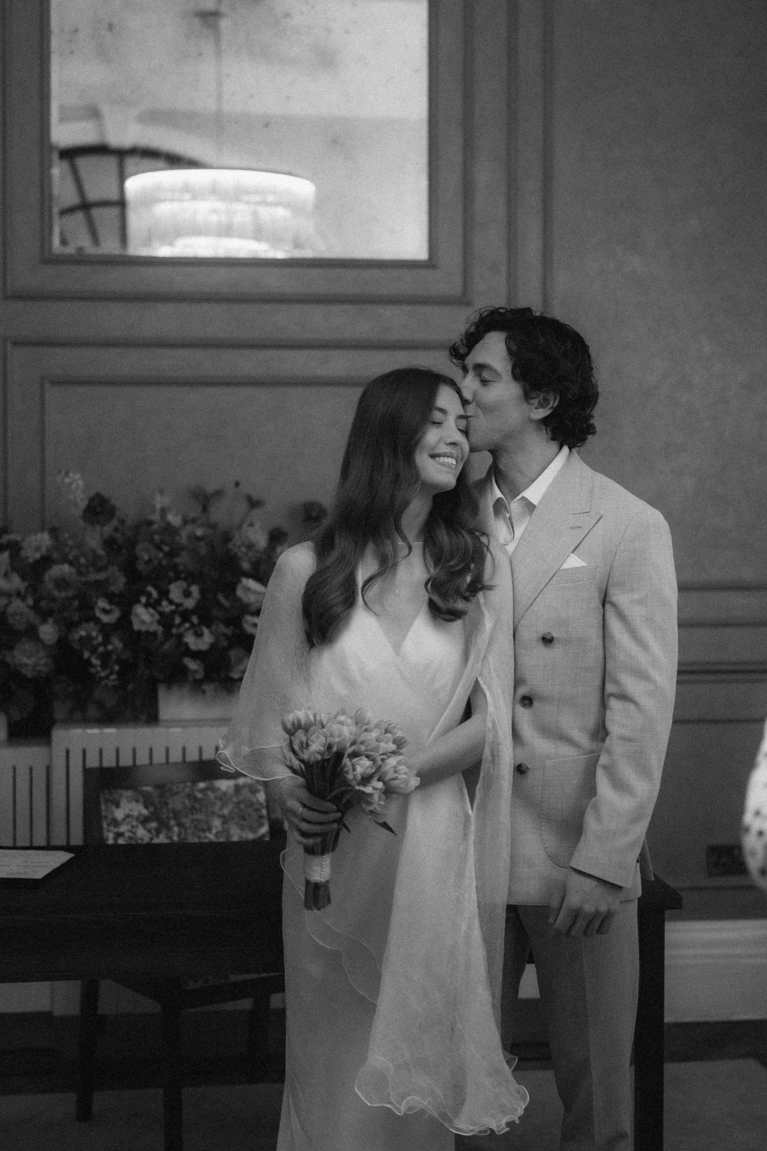 Black and white wedding photo of groom kissing bride on cheek, bride holding bouquet in elegant indoor setting.
