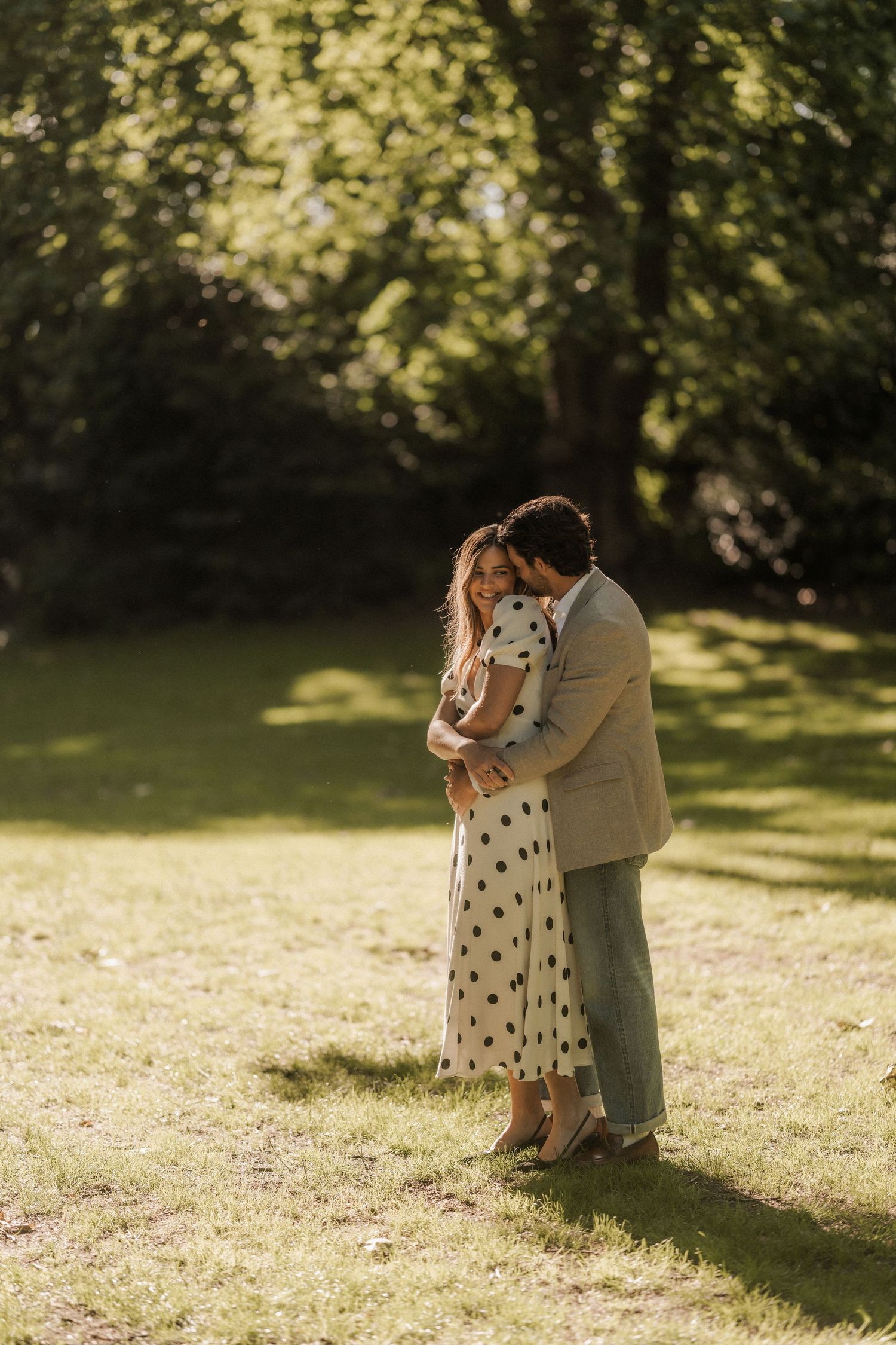 A couple embraces tenderly in a sunlit park, woman in polka dot dress, man in tan blazer.