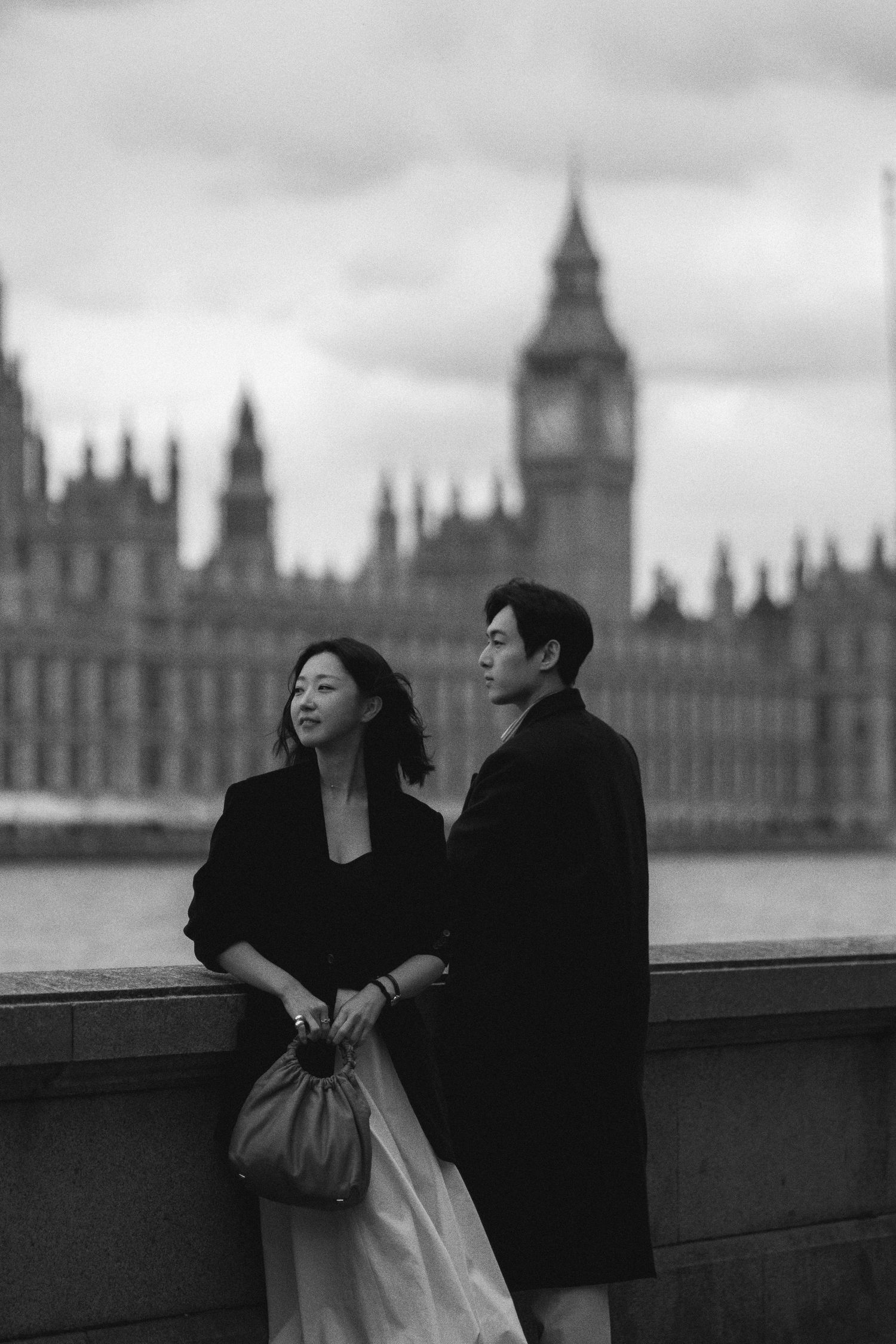 A couple in dark clothing stands along the Thames River with London's Houses of Parliament visible in the background.