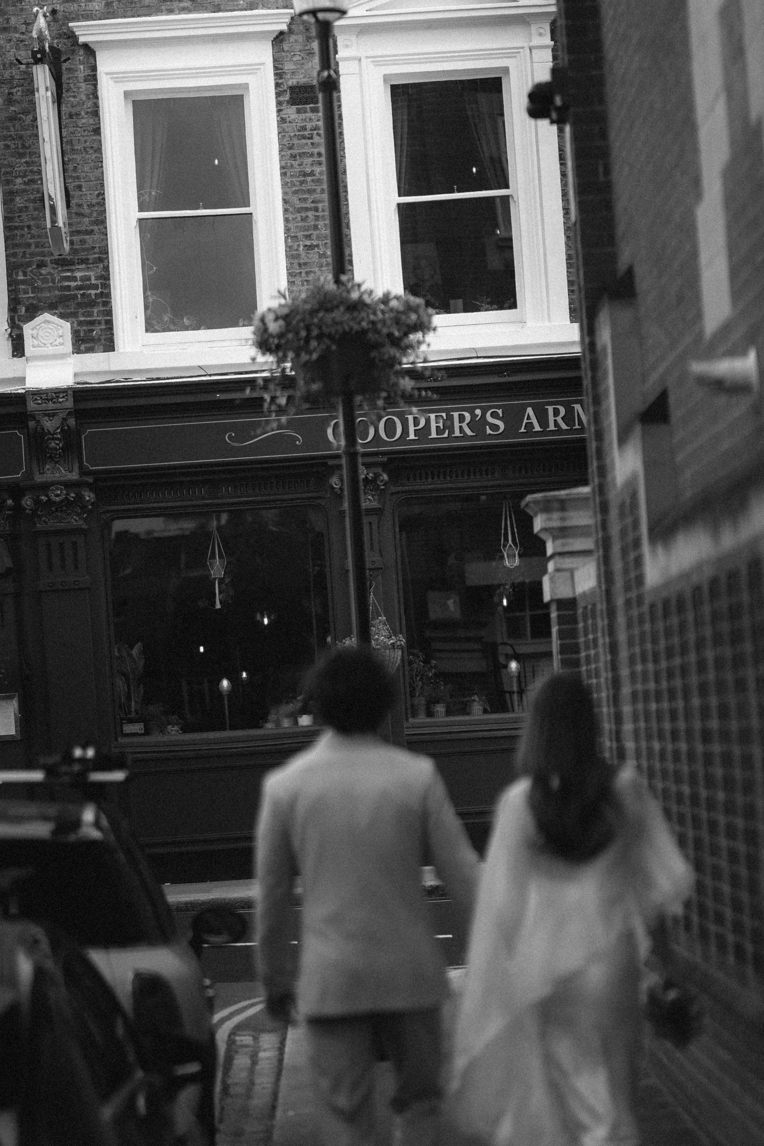 Black and white photo of a couple walking toward Cooper's Arms pub on a cobblestone street in London.