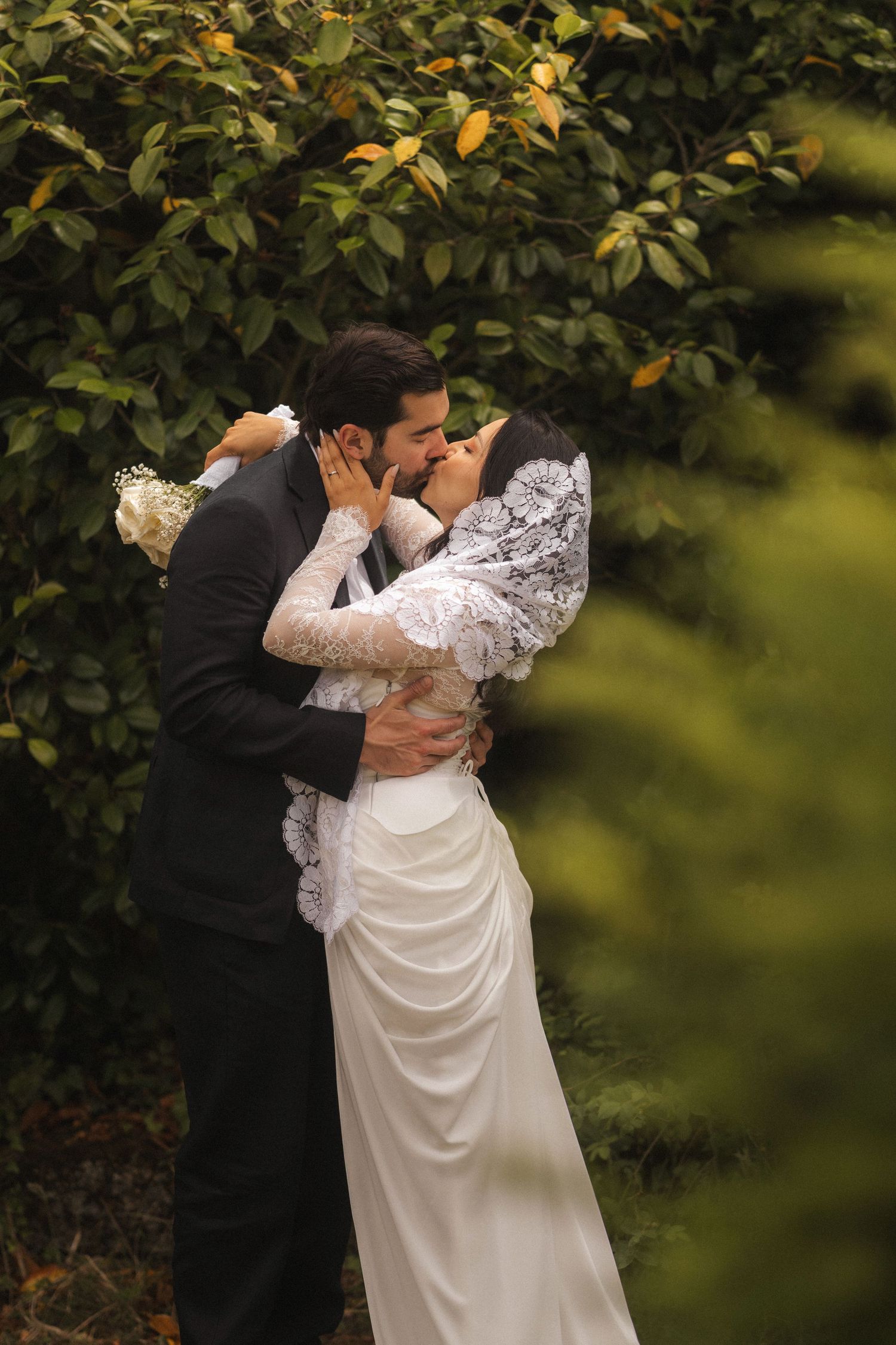 Bride and groom share a romantic kiss surrounded by lush green foliage, bride wearing white lace dress and veil.