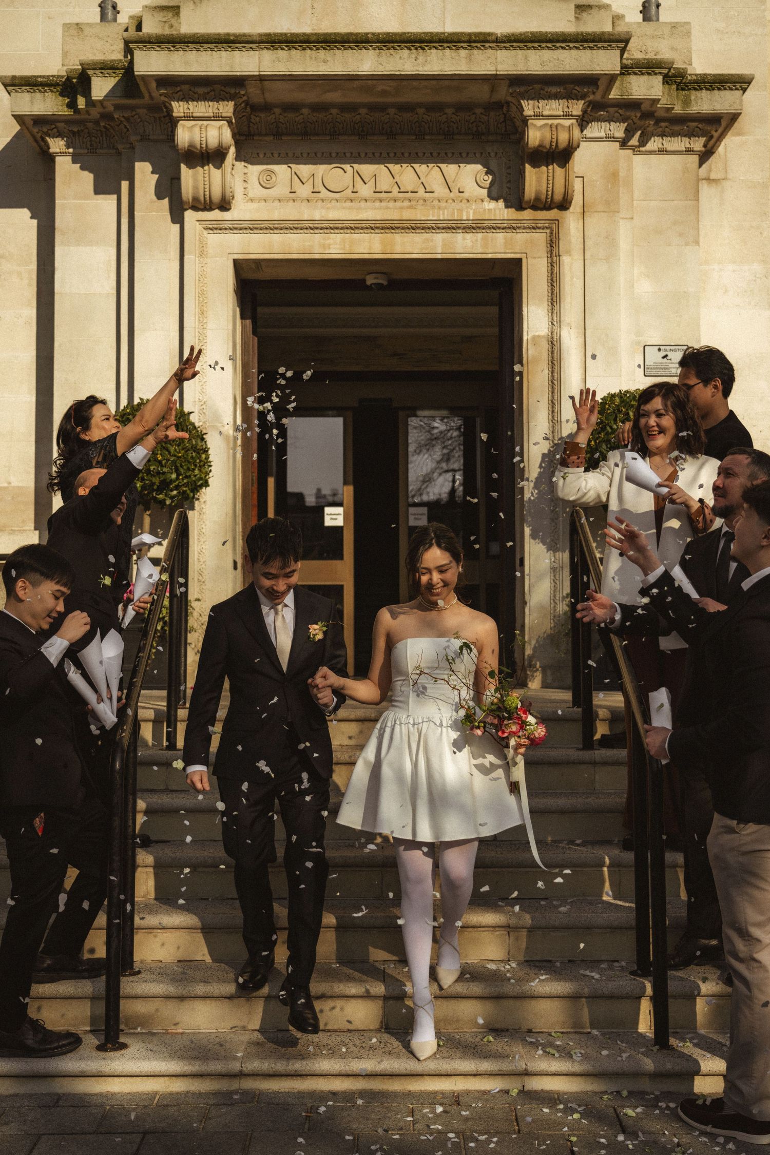 Bride and groom exit a classic stone building as guests toss white confetti during a joyful wedding celebration.