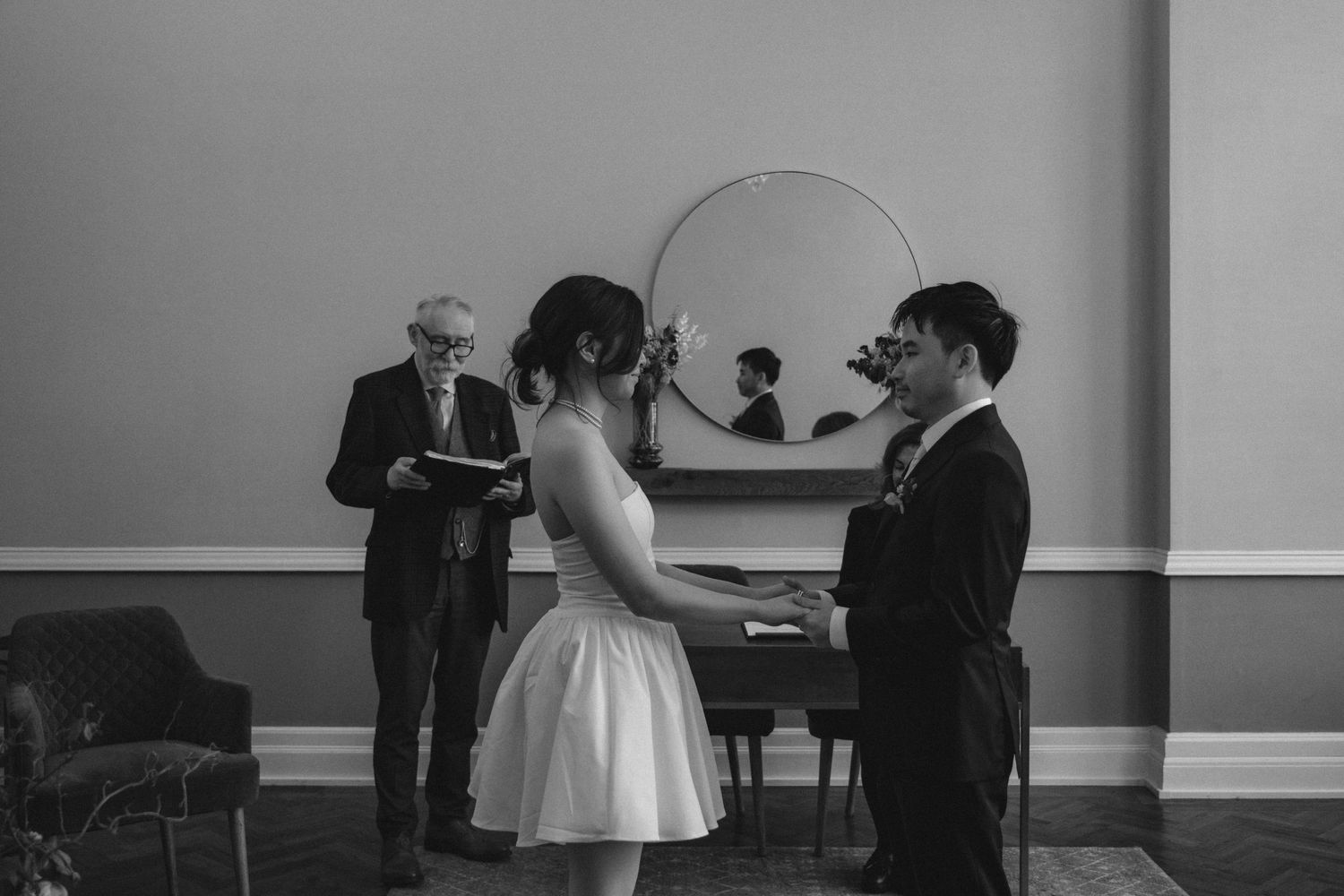 Black and white wedding ceremony scene showing people gathered around a piano in an elegant room.
