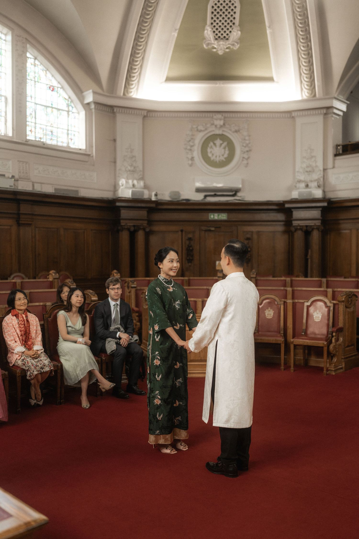 Couple in traditional Vietnamese ao dai attire exchange vows in an elegant wood-paneled ceremony hall with guests seated.