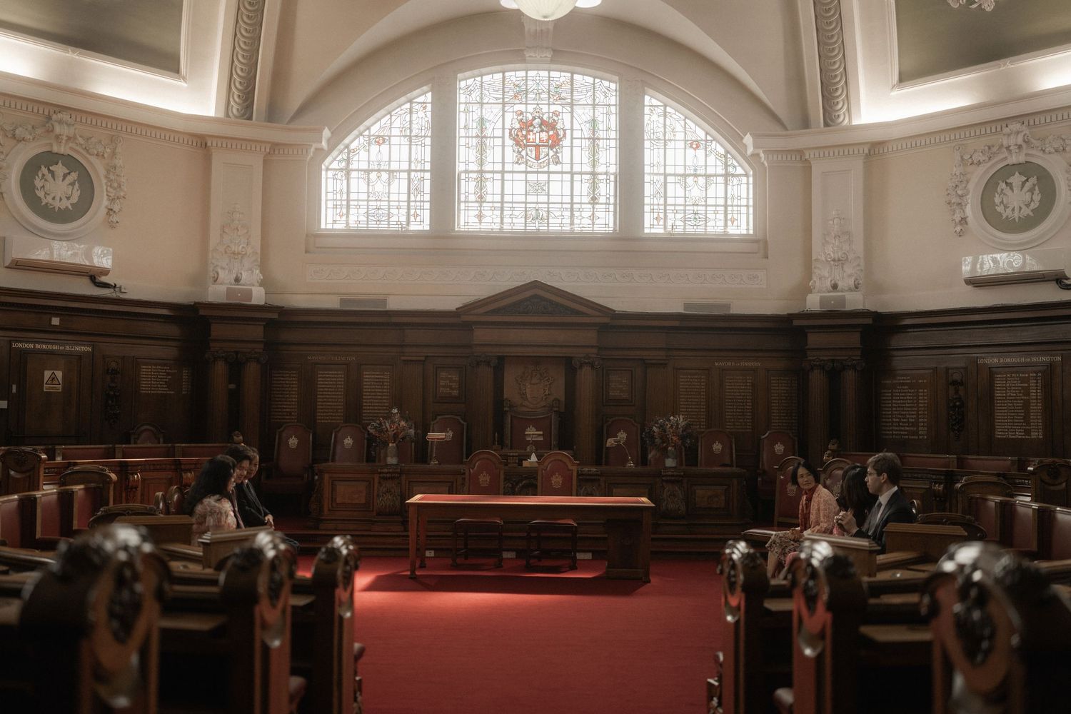 Grand courtroom interior with wooden benches, red carpet, and ornate stained glass window in a historic courthouse.