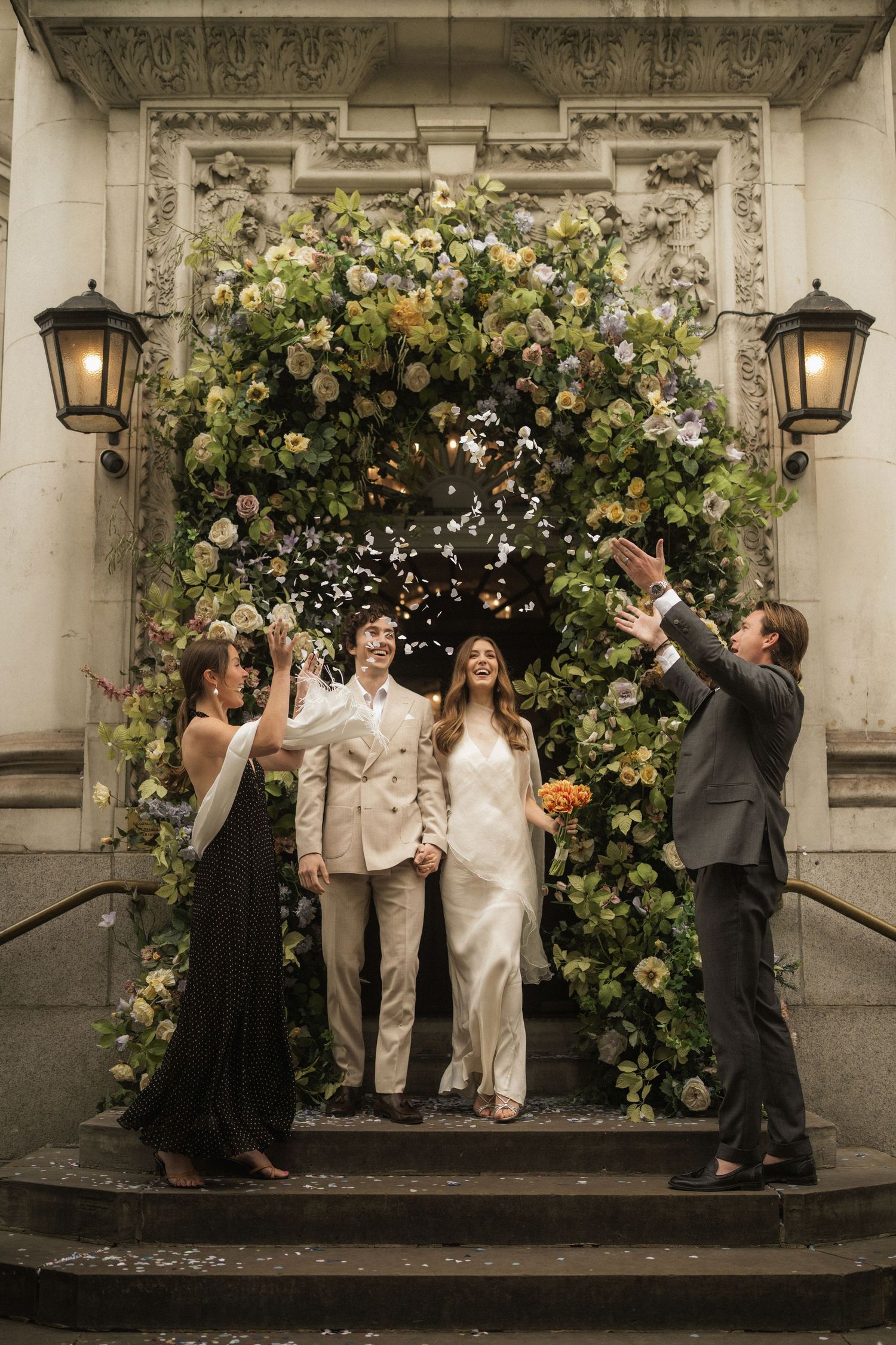 Couple exits floral arch ceremony entrance as guests toss confetti petals at elegant wedding celebration.
