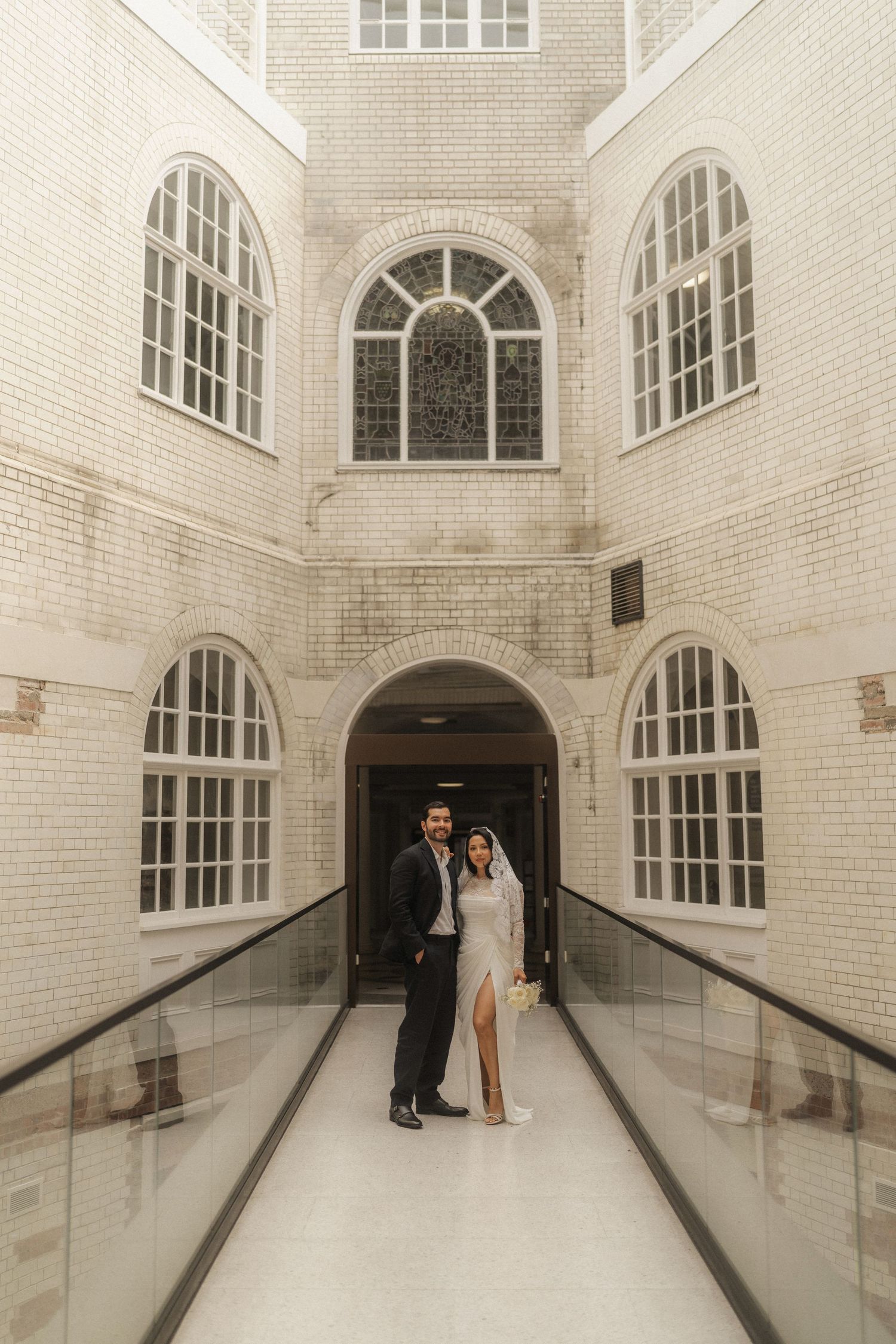 Couple in wedding attire posing in elegant white brick hallway with arched windows and glass railings.