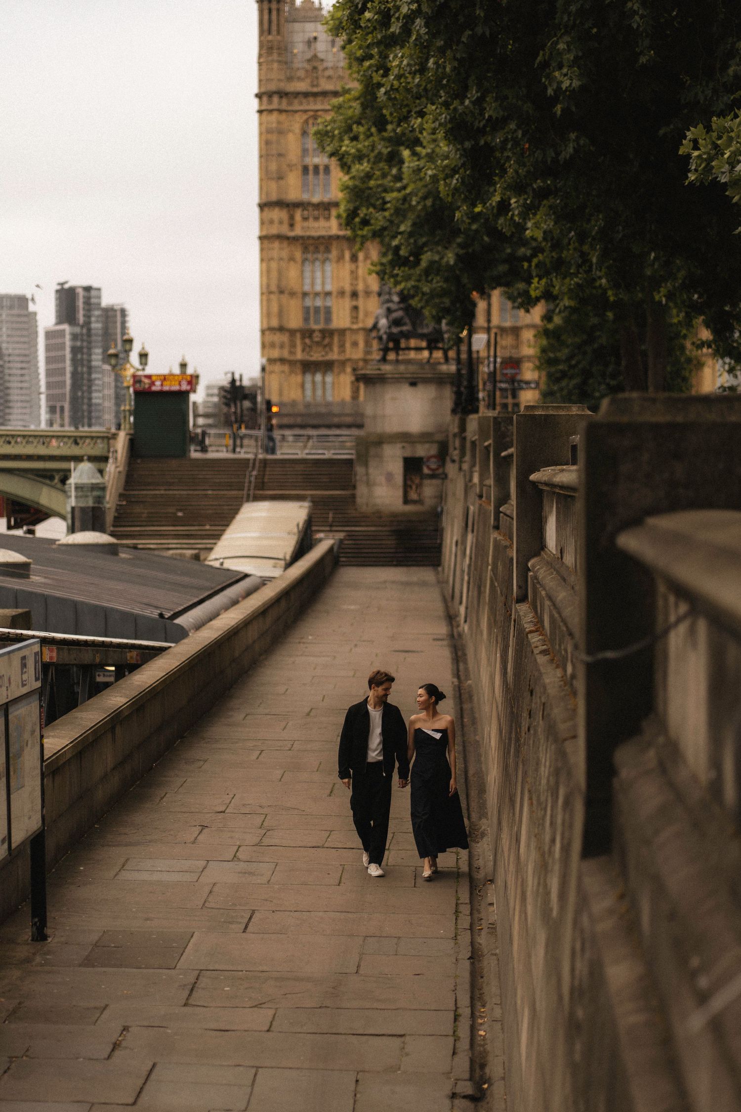 People walking along a stone path bordered by historic buildings and trees in an urban setting.