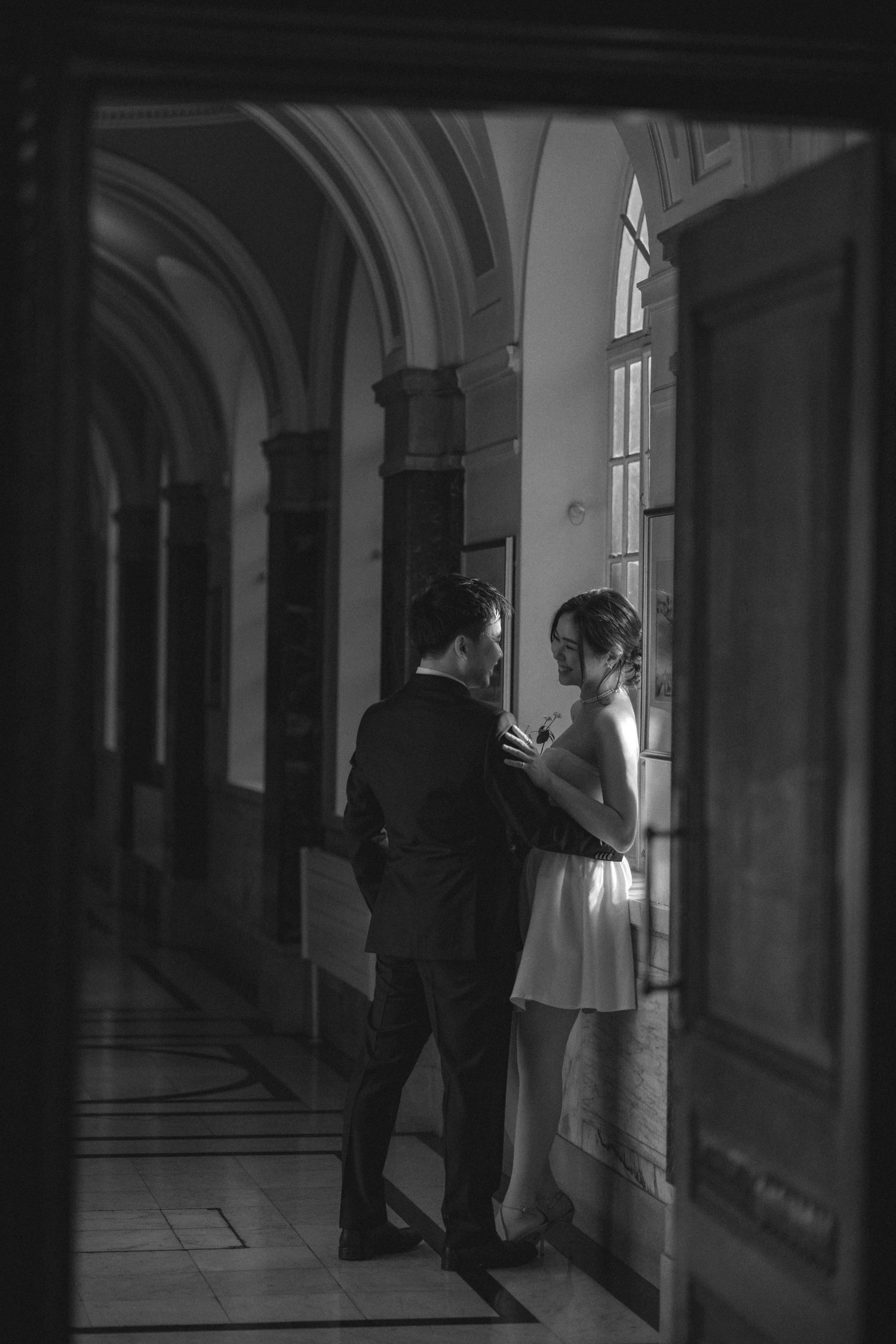 A moody black and white series of figures in a dark corridor with arched windows creating dramatic shadows and silhouettes.
