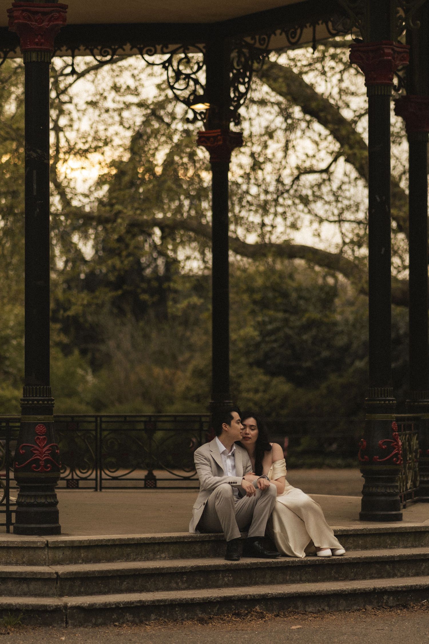A romantic couple sits together on stone steps under an ornate gazebo at sunset in a park setting.