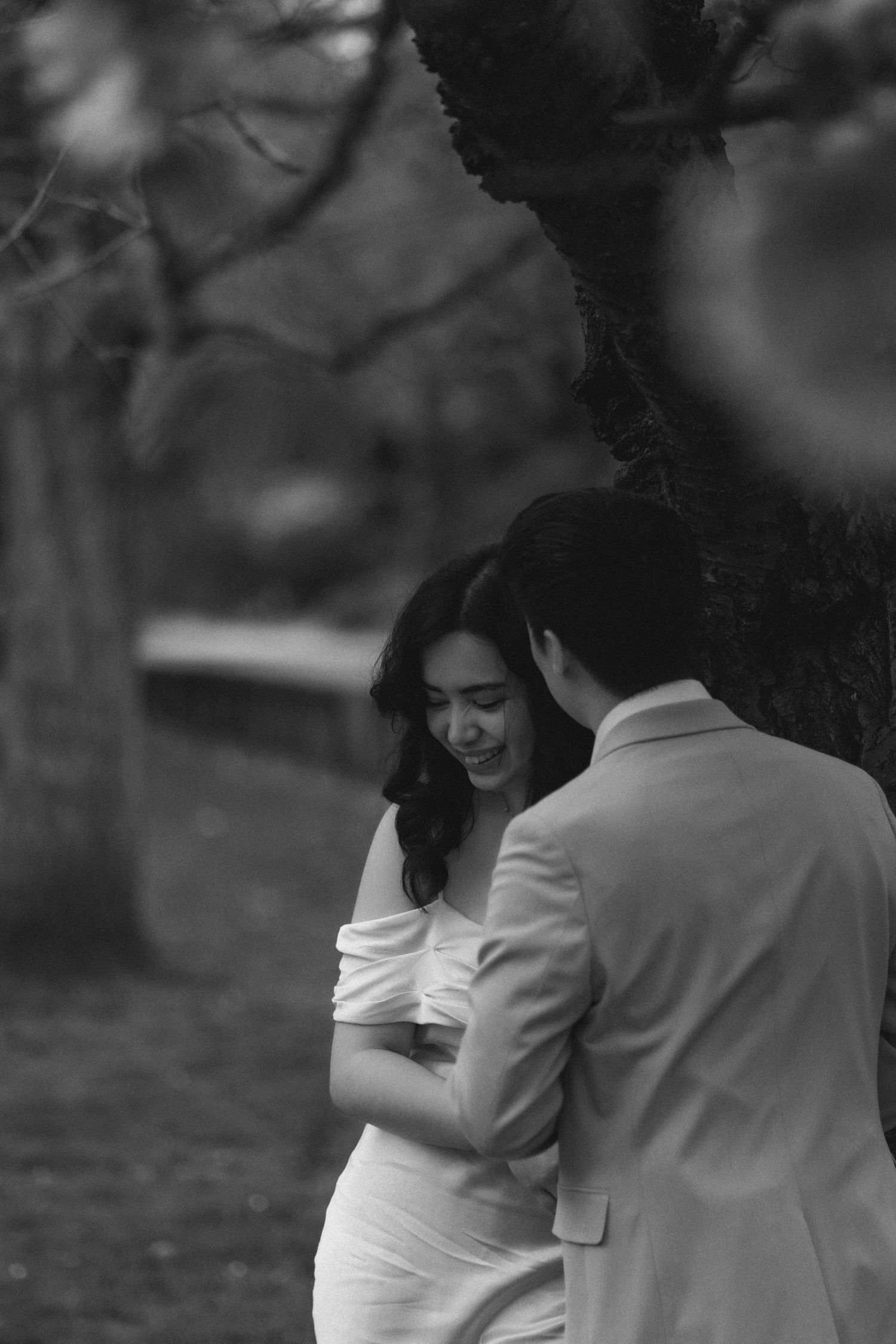 A couple embraces in a romantic black and white photograph taken outdoors in a park setting.