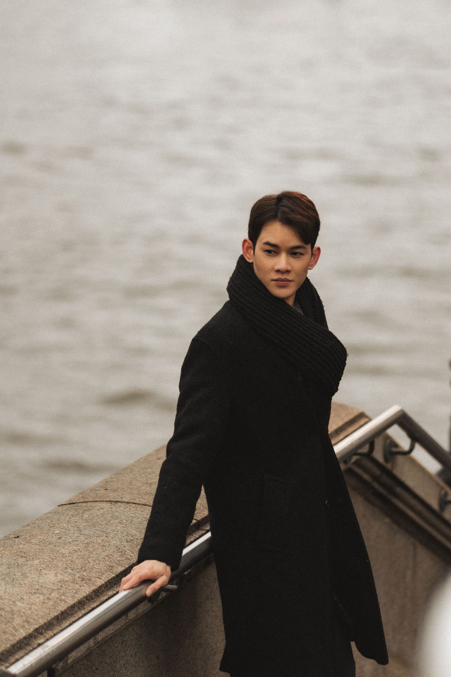 Person in a black coat stands near a metal railing by the water on an overcast day.