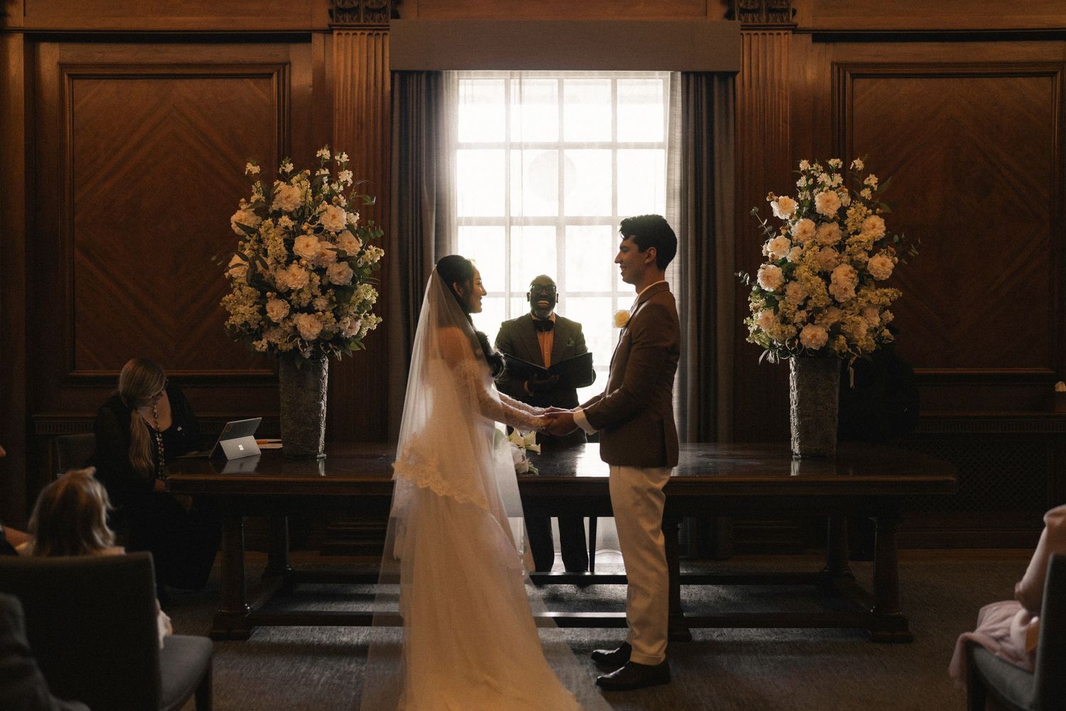 A wedding ceremony takes place in a grand dark wood-paneled room with elegant floral arrangements and natural light.