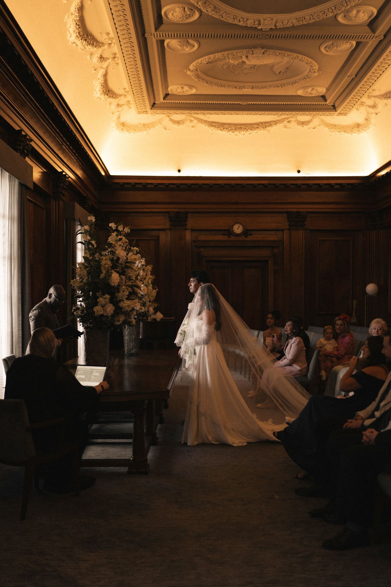 A bride in a flowing white gown and long veil walks down the aisle in an elegant wood-paneled room with ornate ceiling details.
