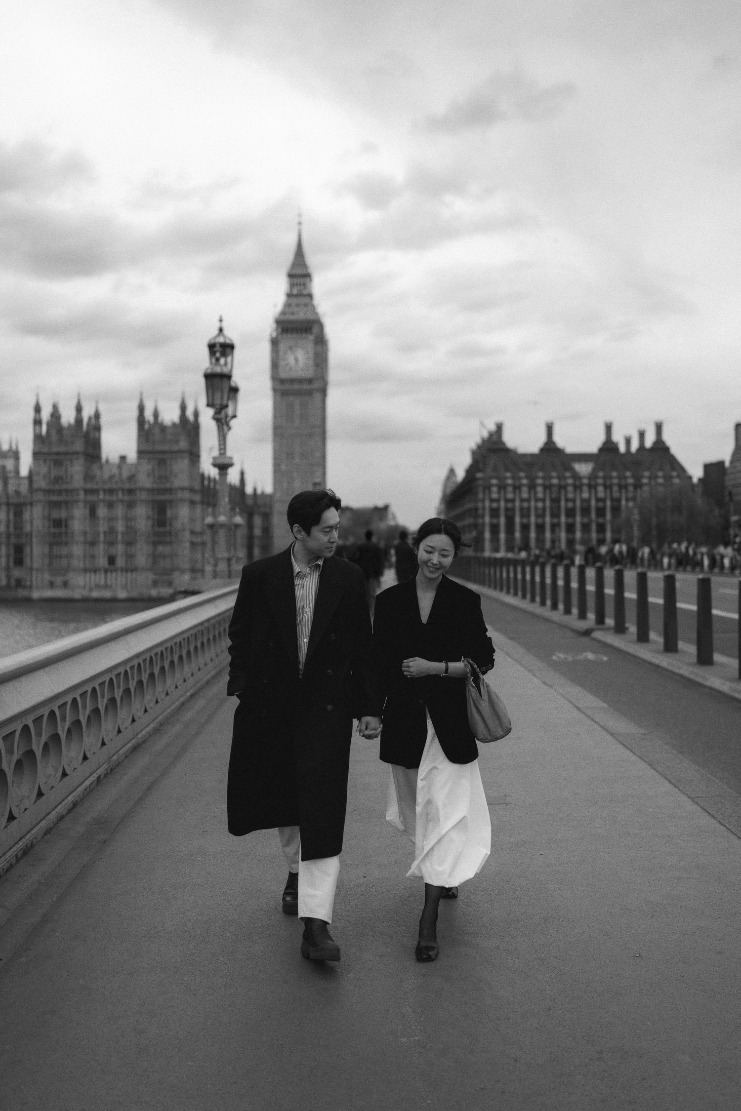 A couple walks across Westminster Bridge in London with Big Ben and Parliament buildings in the background, black and white photo.