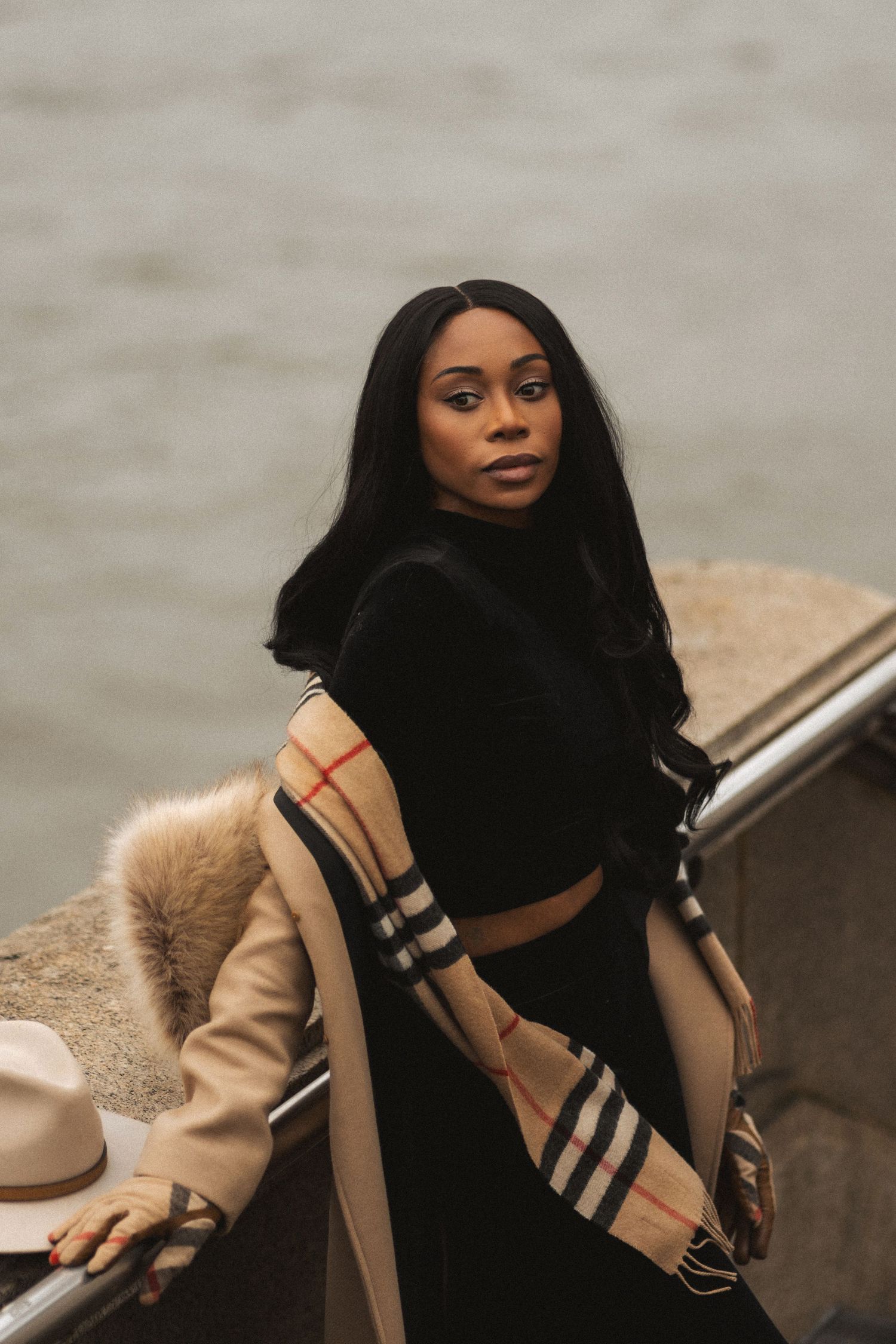A person in a black turtleneck and beige coat with fur trim poses outdoors on an overcast day.