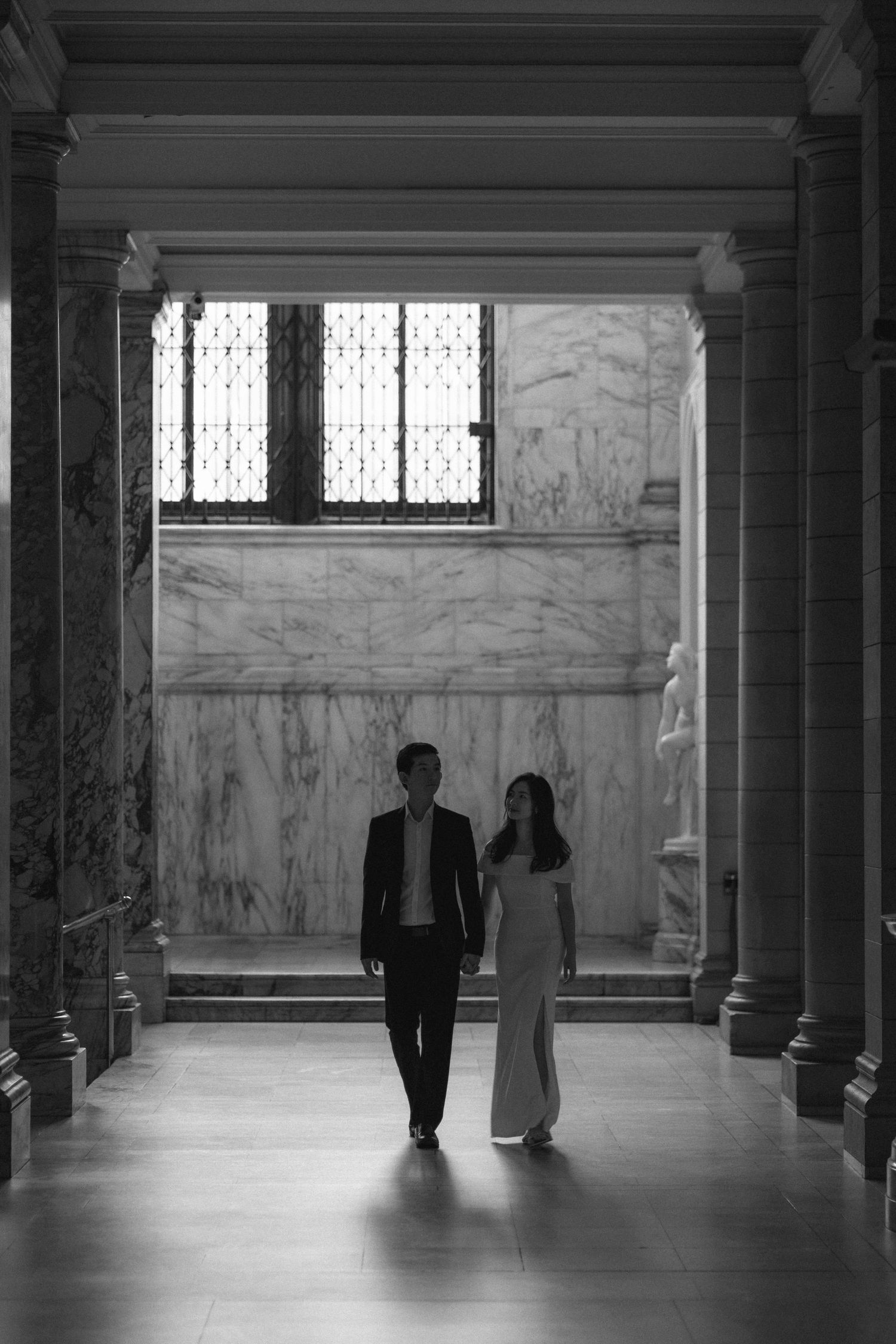 Two people stand in silhouette against marble walls in a grand architectural space with columns and high windows.