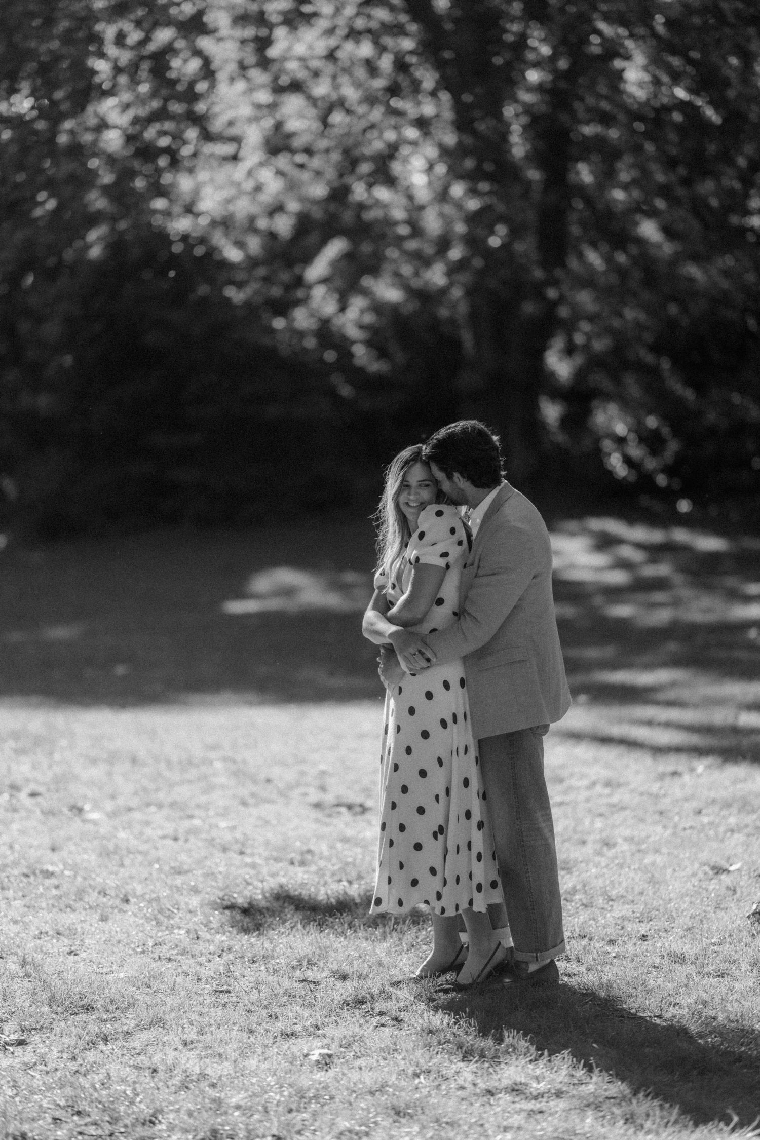 Black and white photo of a couple embracing tenderly in a sunlit park, woman in polka dot dress.