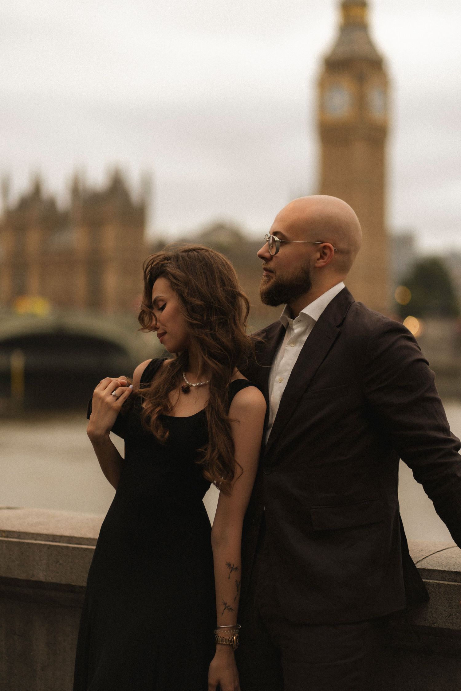 Elegant couple in black attire posing near the Thames with Big Ben in the background in London.