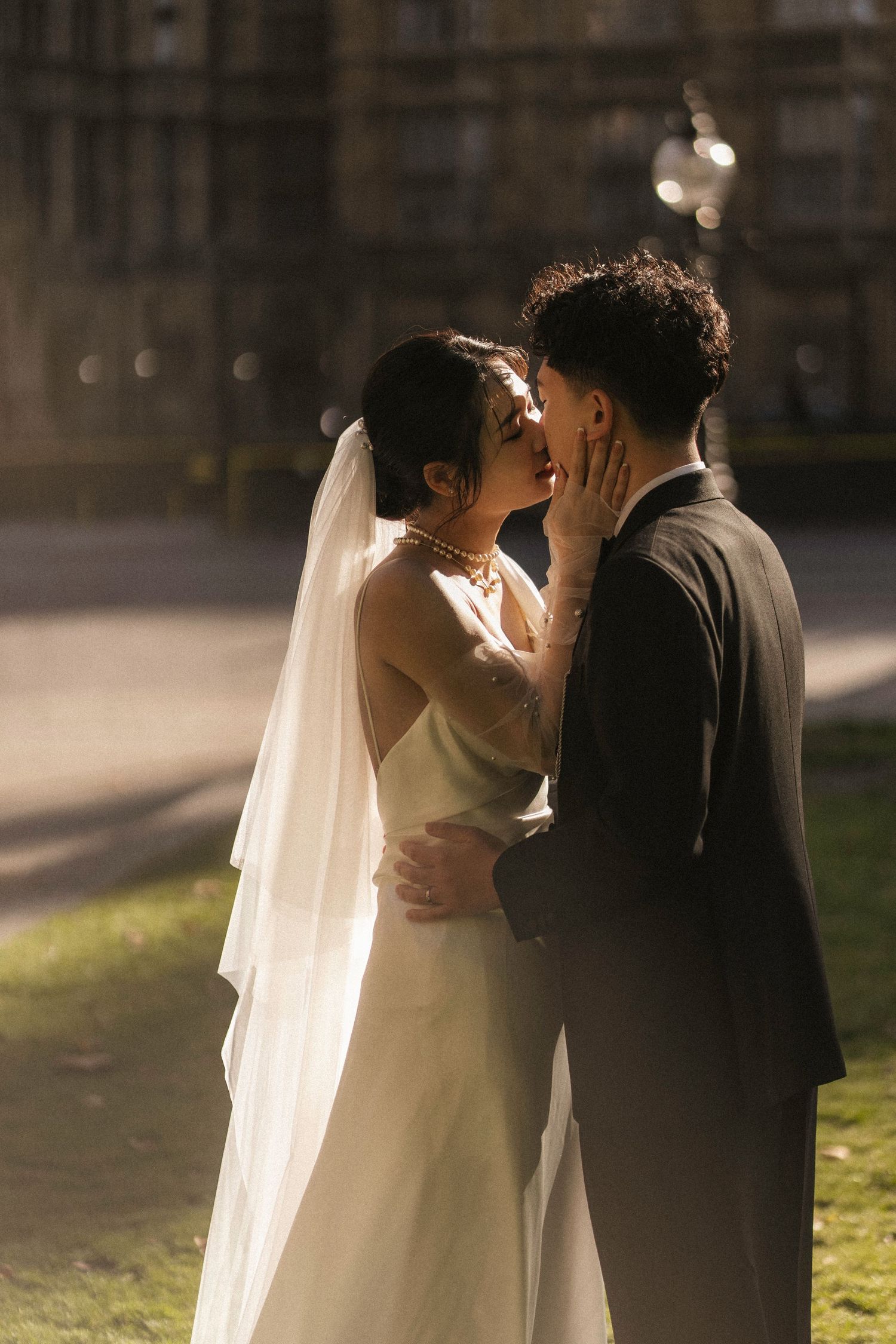 A bride and groom share a romantic kiss outdoors, she wears a white gown with veil, he wears a dark suit.