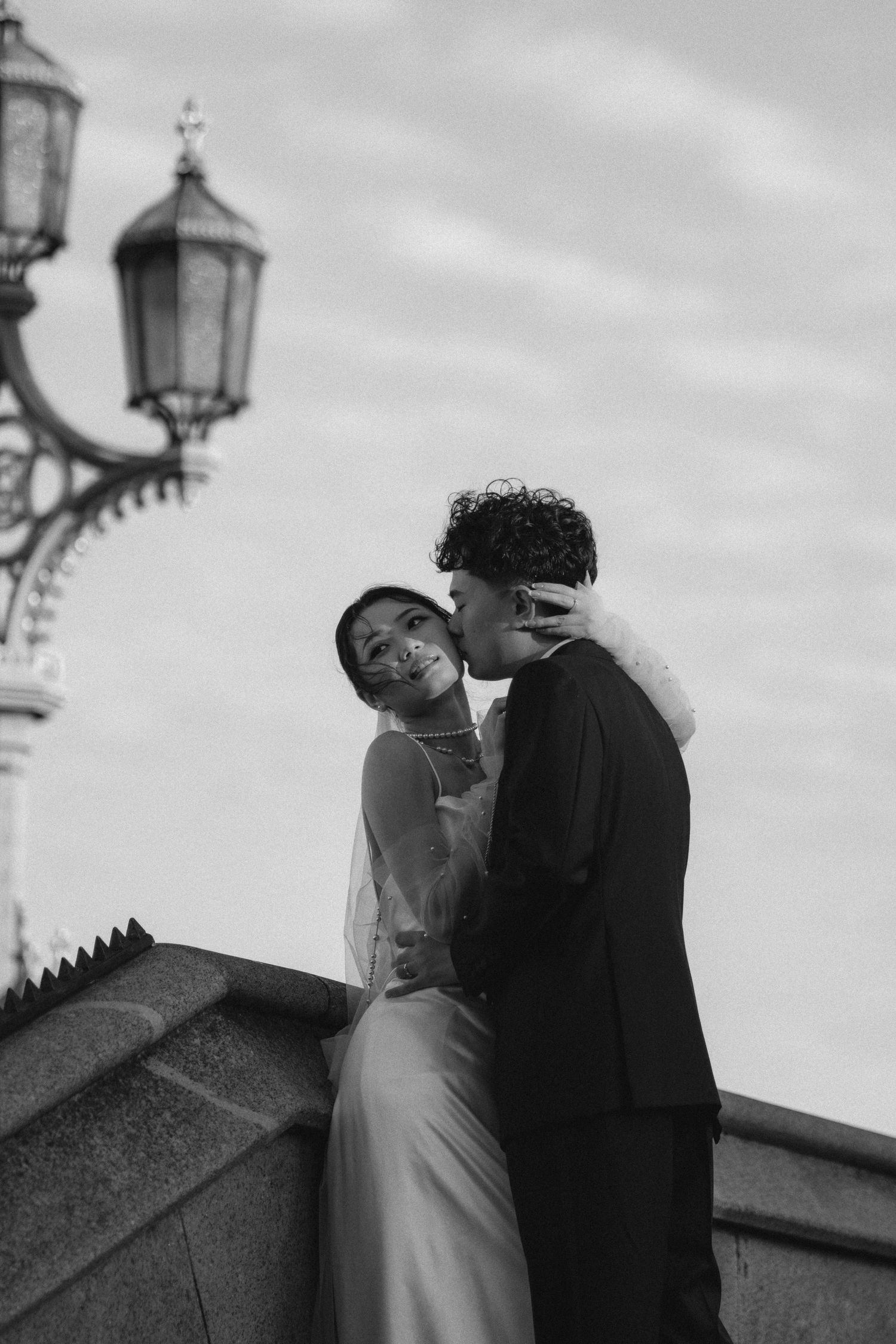 Black and white photo of a couple in wedding attire sharing a romantic moment near a vintage street lamp.