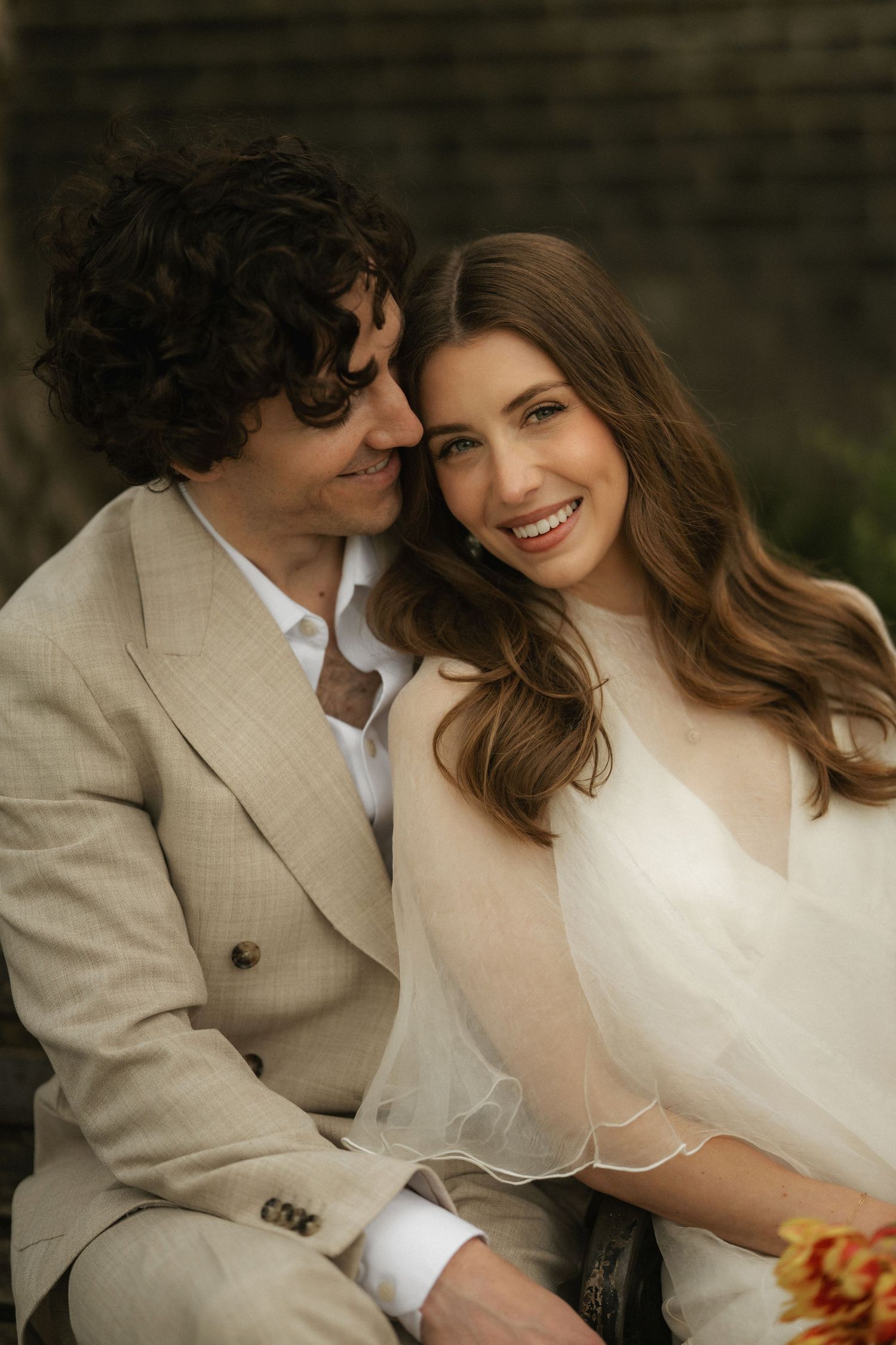 A smiling bride and groom in elegant wedding attire sit closely together, the groom in a beige suit nuzzling the bride.
